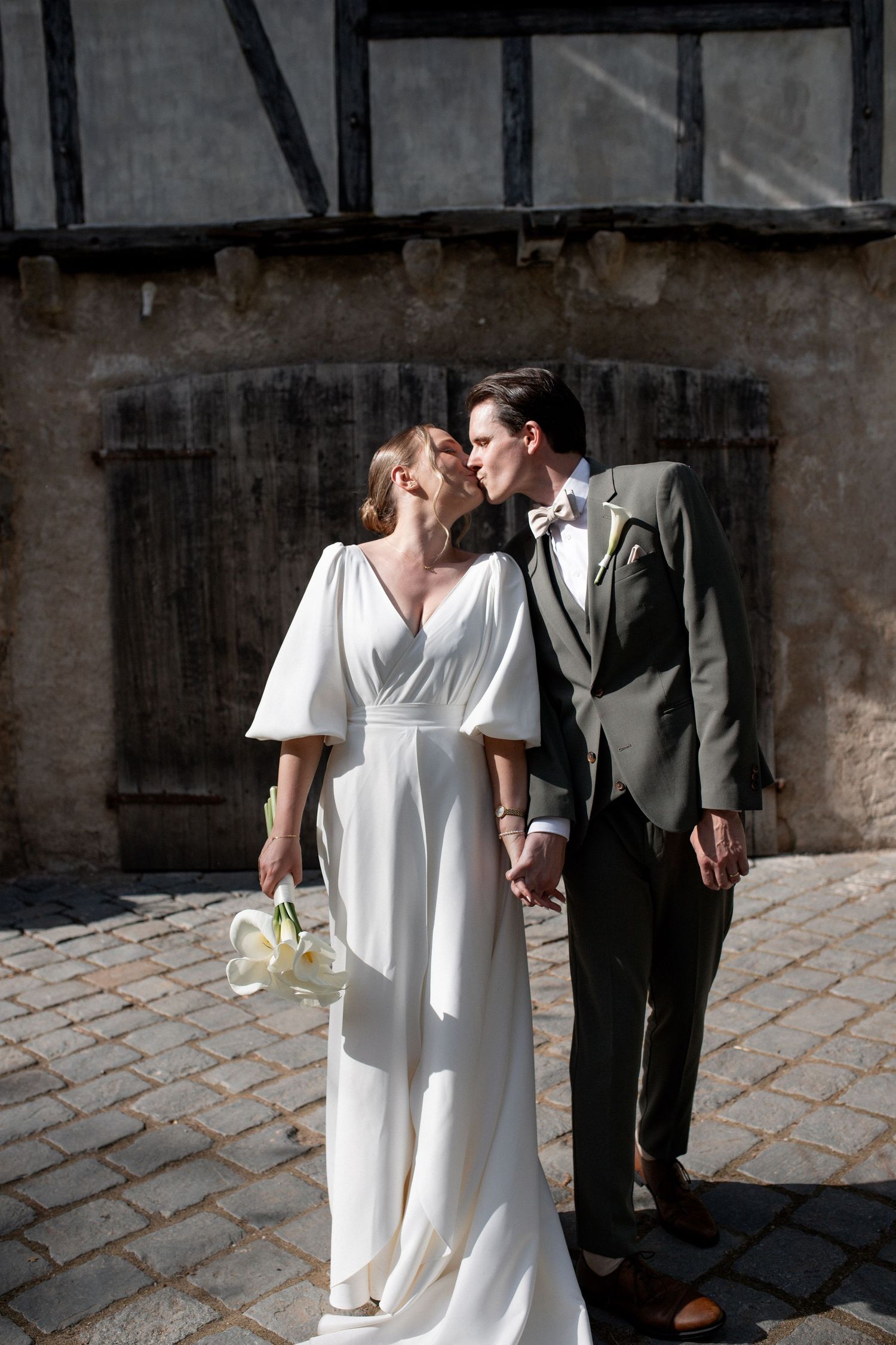 Bride in white flutter-sleeve gown and groom in olive suit share a romantic kiss on cobblestone street.