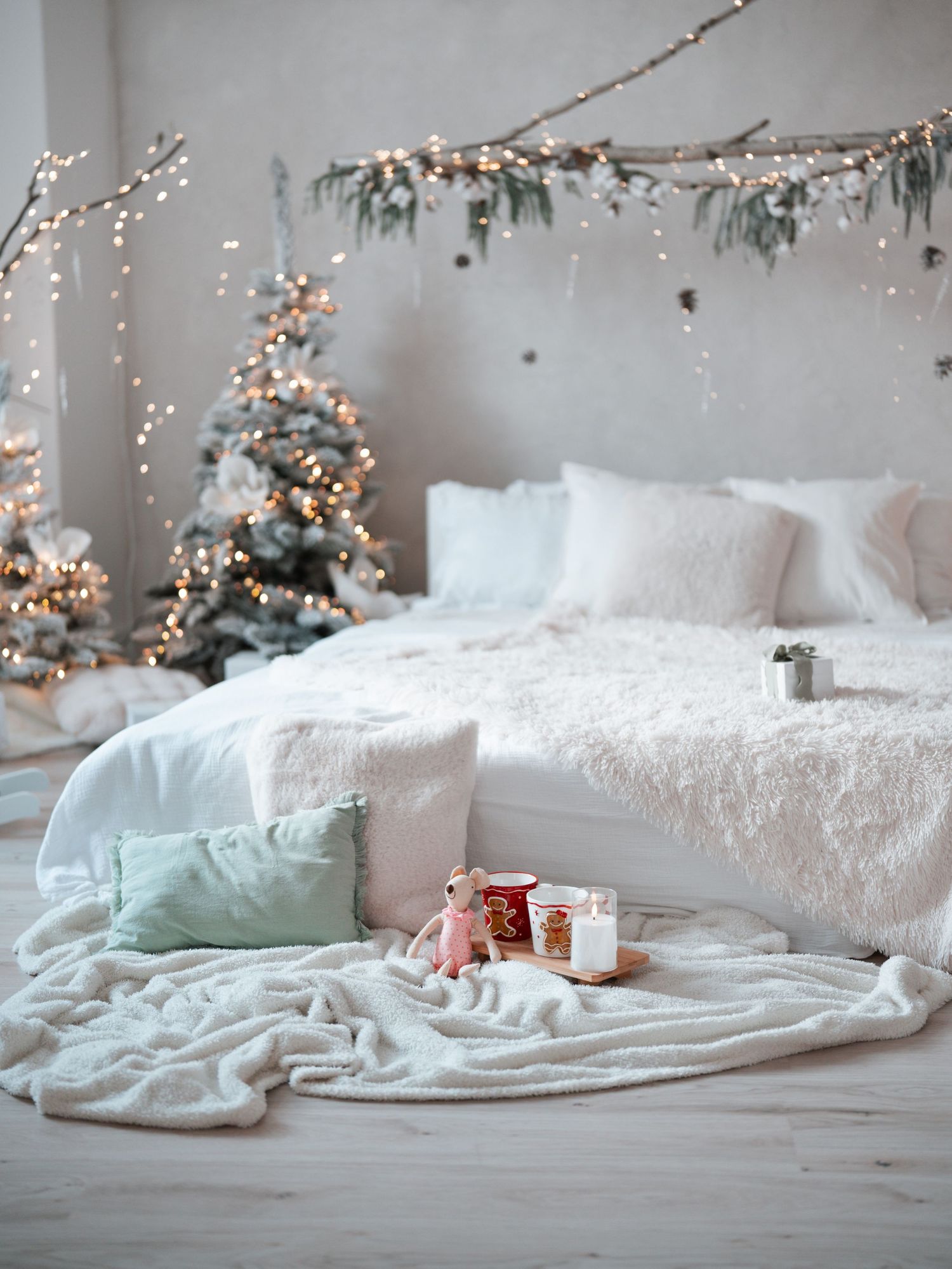 Cozy white bedroom with Christmas tree, fairy lights, fluffy bedding, and holiday decorations on the floor.