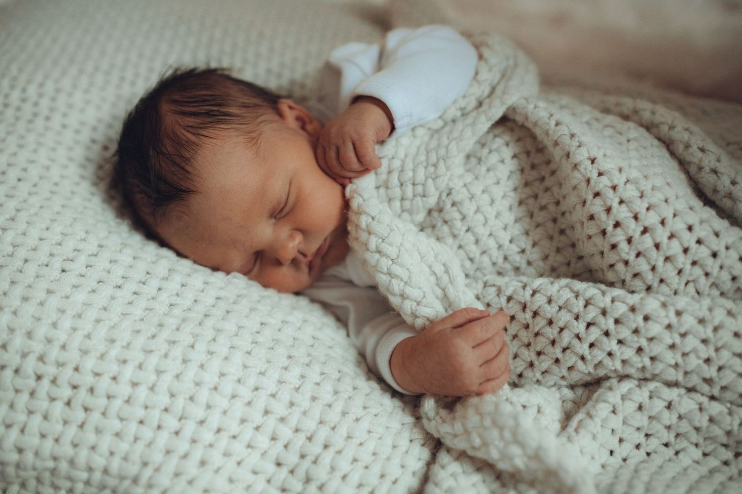 A sleeping newborn baby wrapped in a cozy white knit blanket rests peacefully on soft bedding.