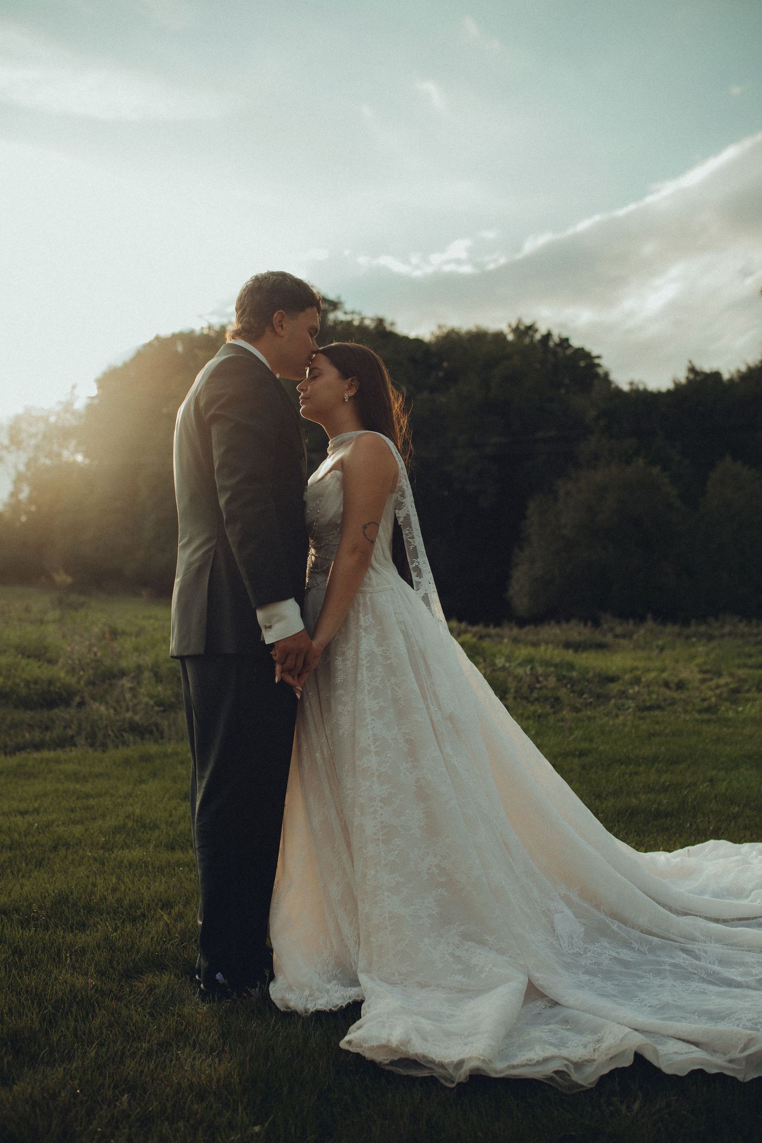 Bride and groom share a romantic moment at sunset in a lush green field, bride wearing a flowing white gown.