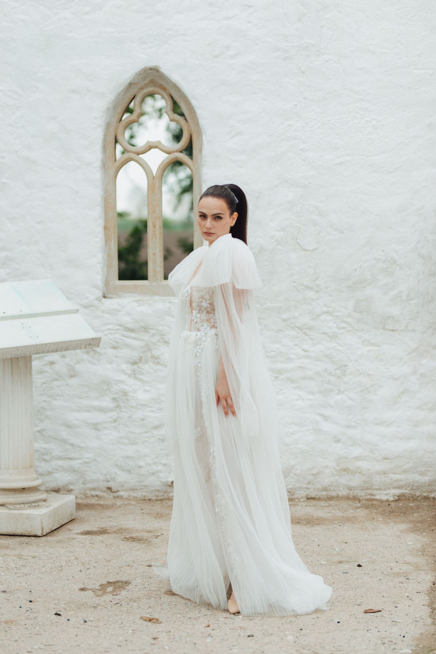 Elegant bridal pose in front of white stone wall with arched windows creates romantic ambiance.