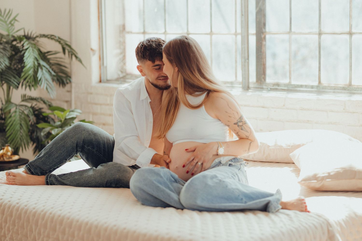 Intimate maternity moment on white bed in sunlit room with modern decor.