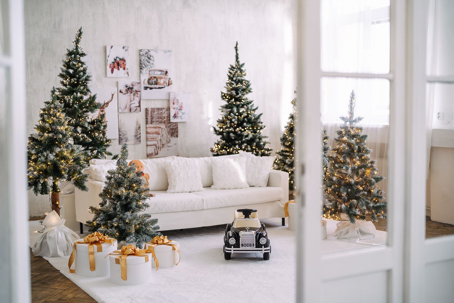 Bright white modern living room decorated with multiple Christmas trees and festive holiday decor along large windows.