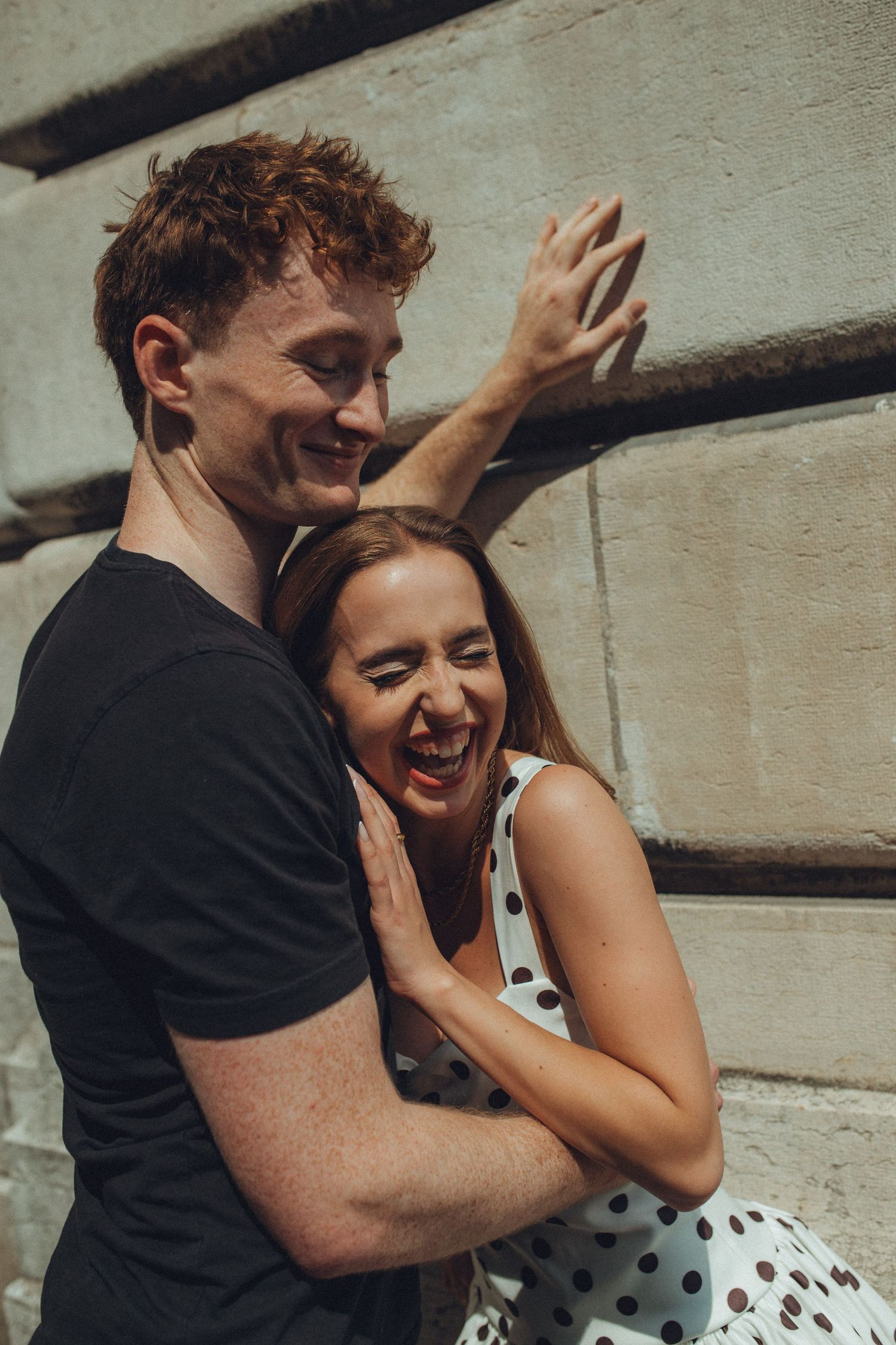A joyful couple shares laughter and embraces against a concrete wall background.
