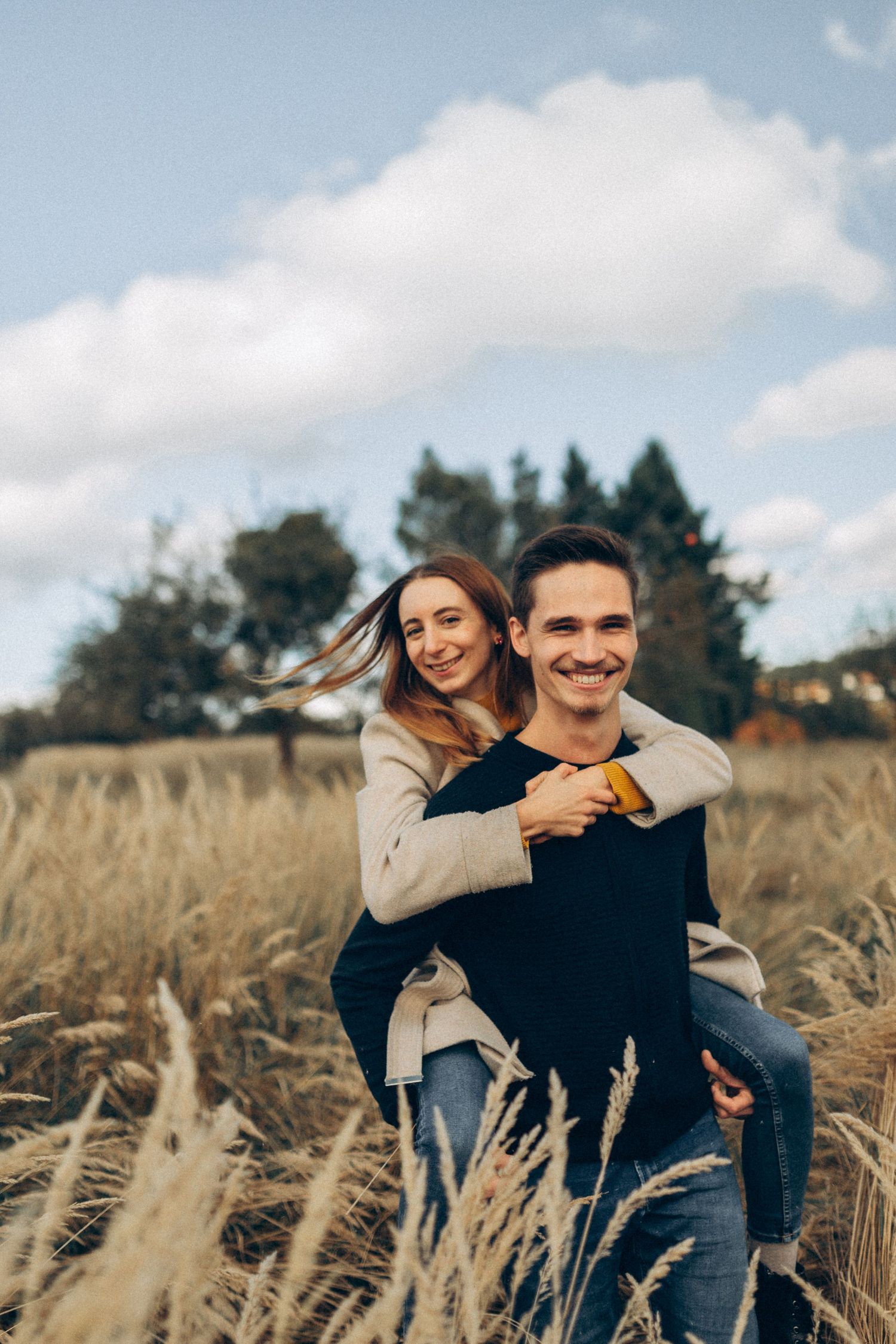 A couple plays and laughs together in a golden field with evergreen trees in the background.