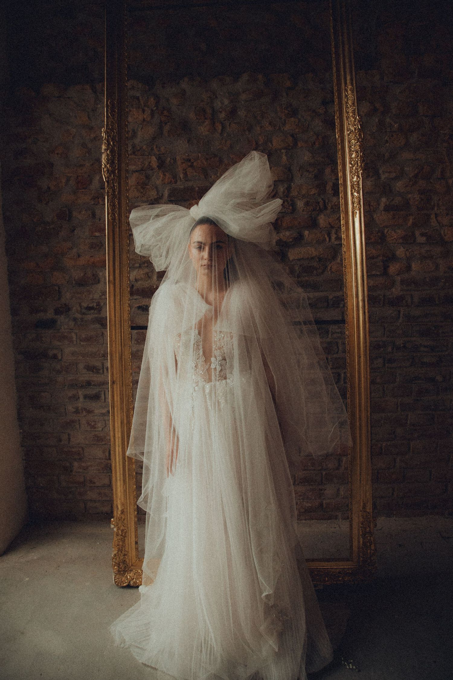 Dramatic bridal portrait in ethereal white gown against dark brick wall with antique gold mirror.