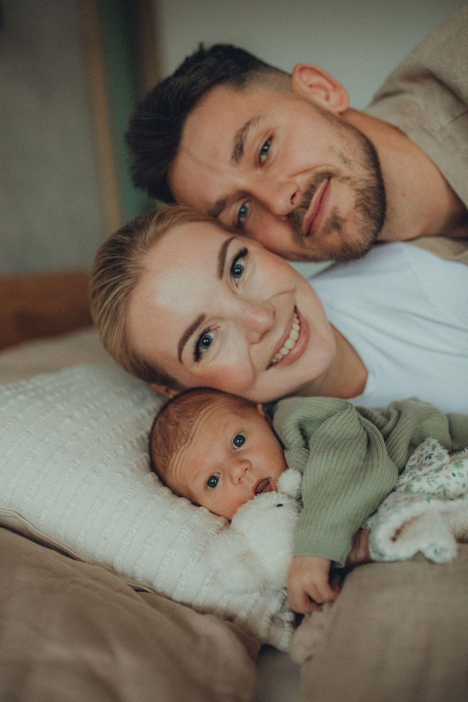 A cozy family portrait showing parents lying down with their newborn baby in a sage green outfit.