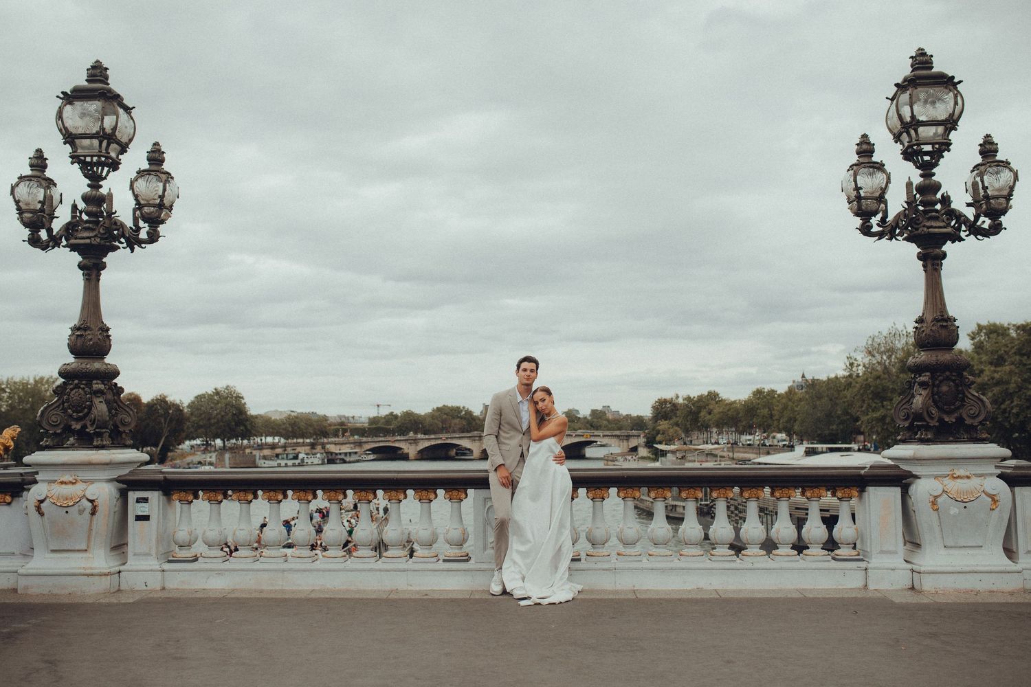 Bride and groom posing on Pont Alexandre III bridge in Paris with ornate lampposts and cloudy sky.