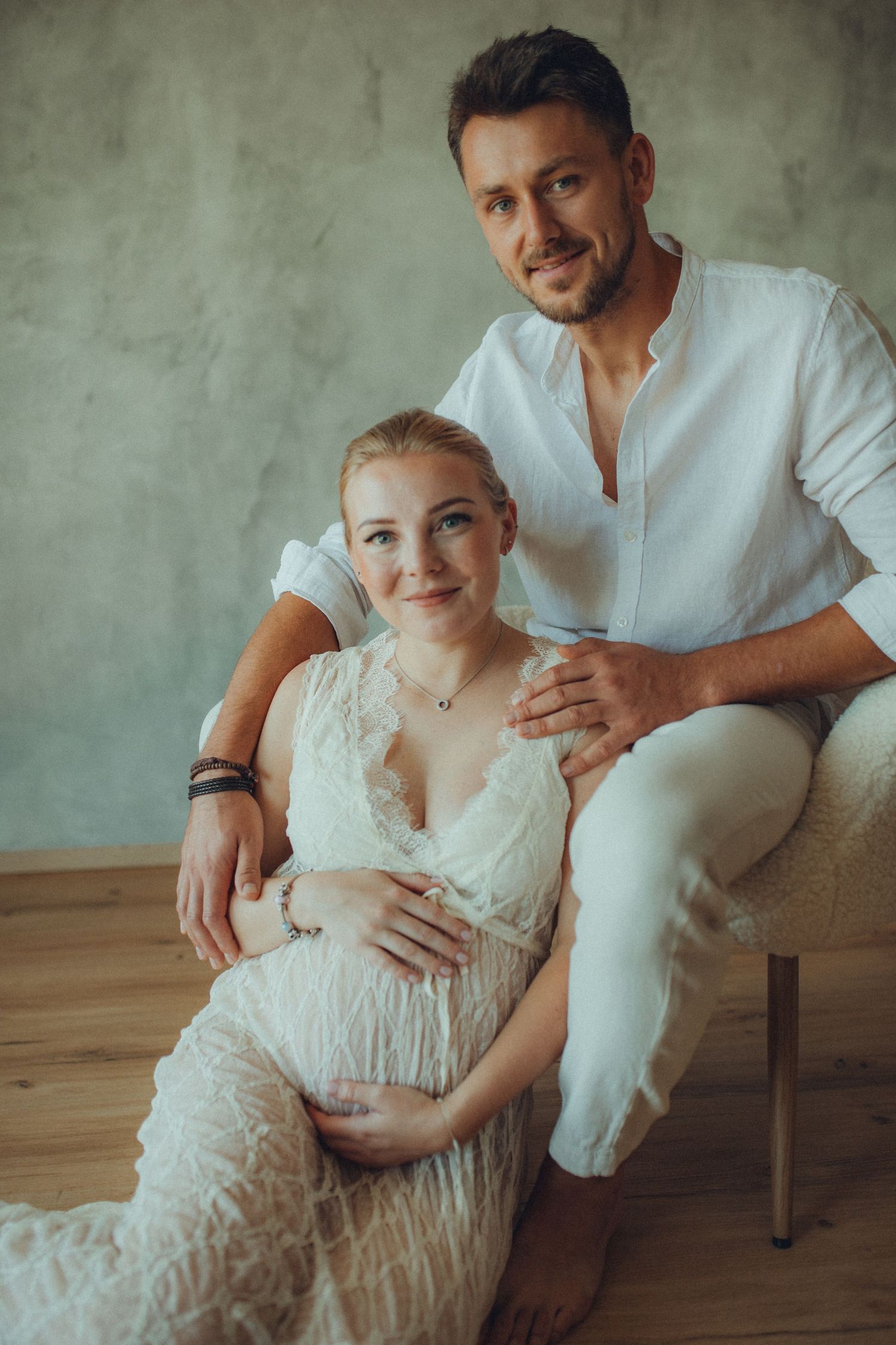 Intimate maternity photo series showing couple in white clothing sitting together on wooden floor against grey wall.