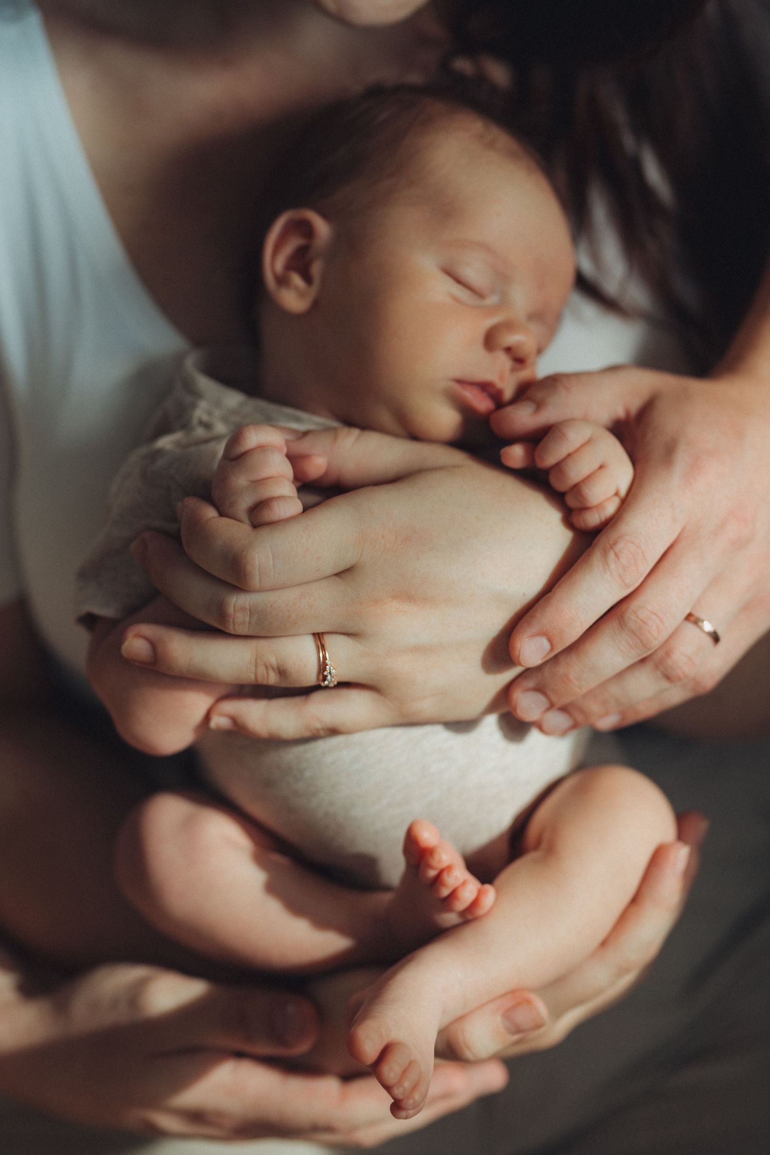 Newborn baby sleeping peacefully in parents hands, mother wearing gold wedding ring, tender family moment.