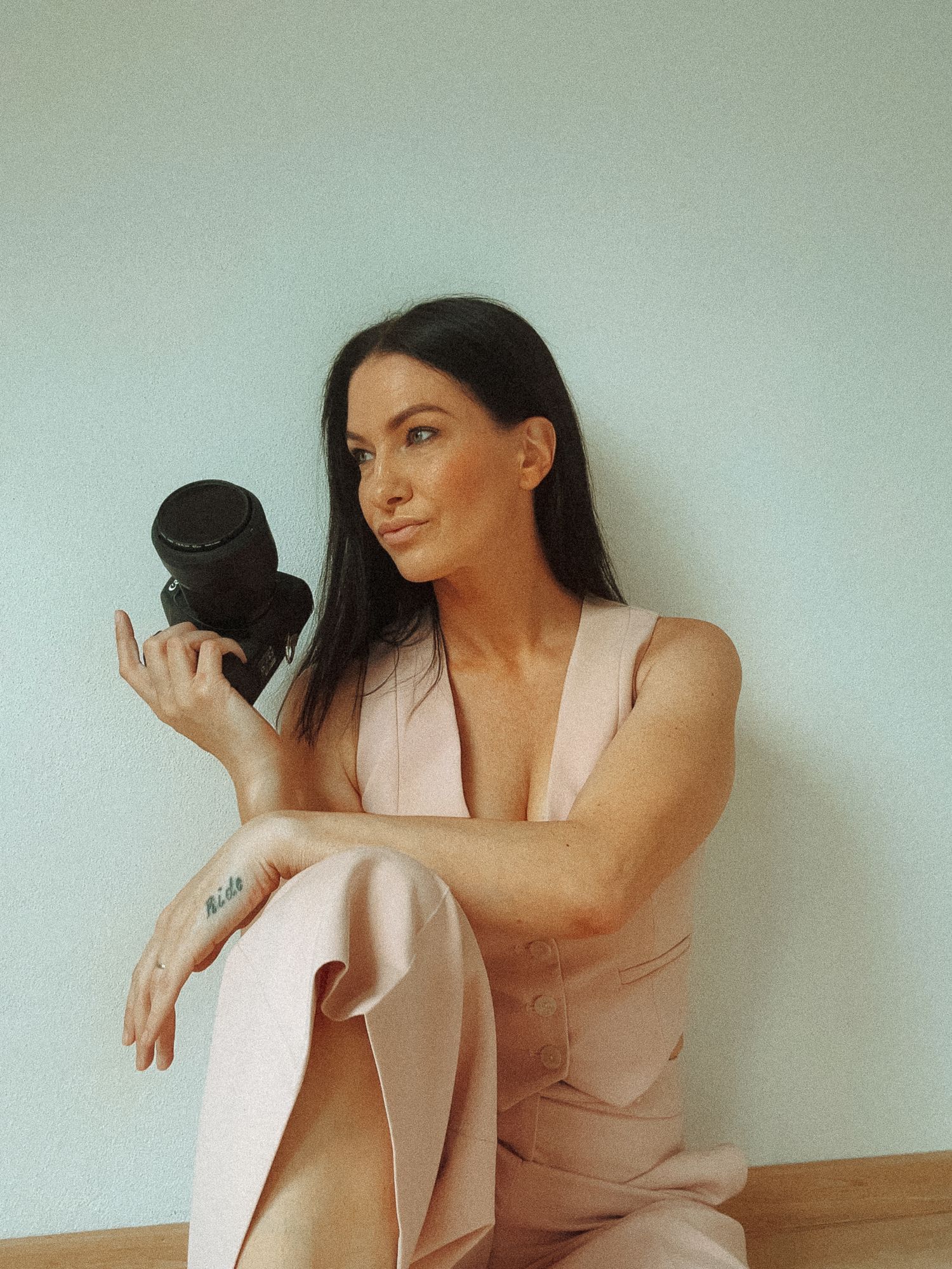 Contemporary photographer poses with vintage camera in muted pink and green tones while sitting on wooden bench.