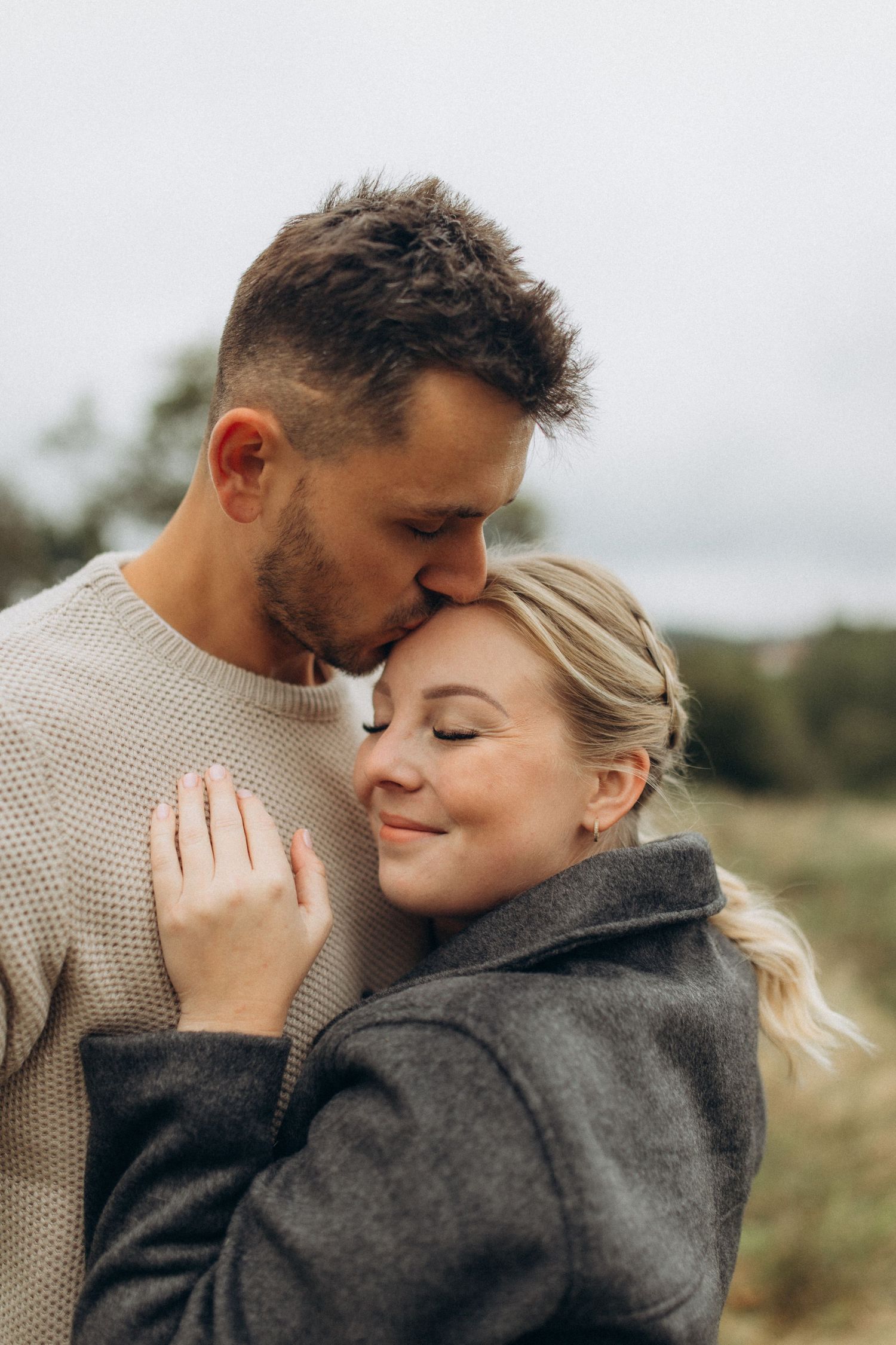 An intimate couple shares a tender moment while wearing cozy sweaters in a natural setting.