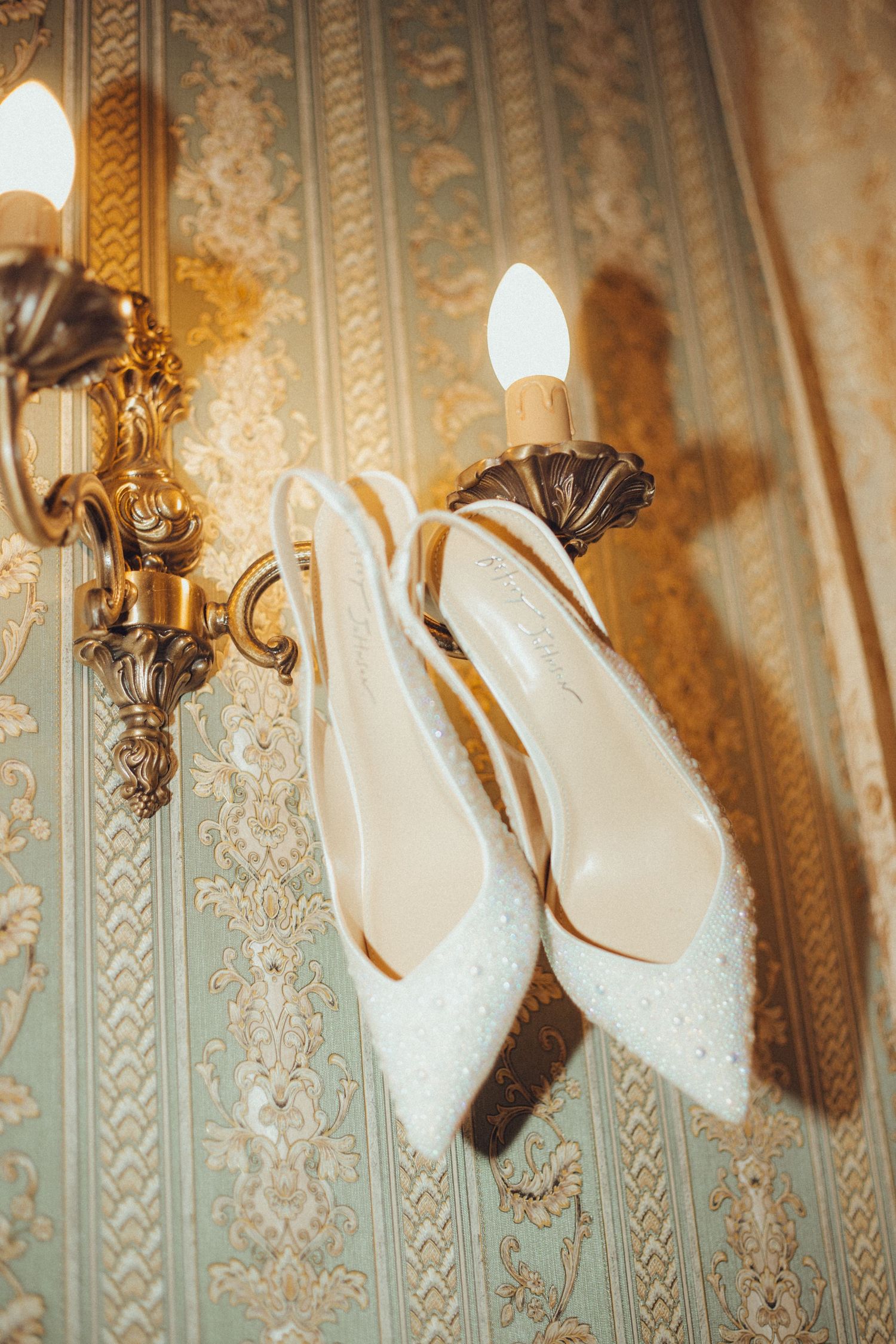 Elegant white bridal shoes with crystal details hanging on ornate wall sconce.