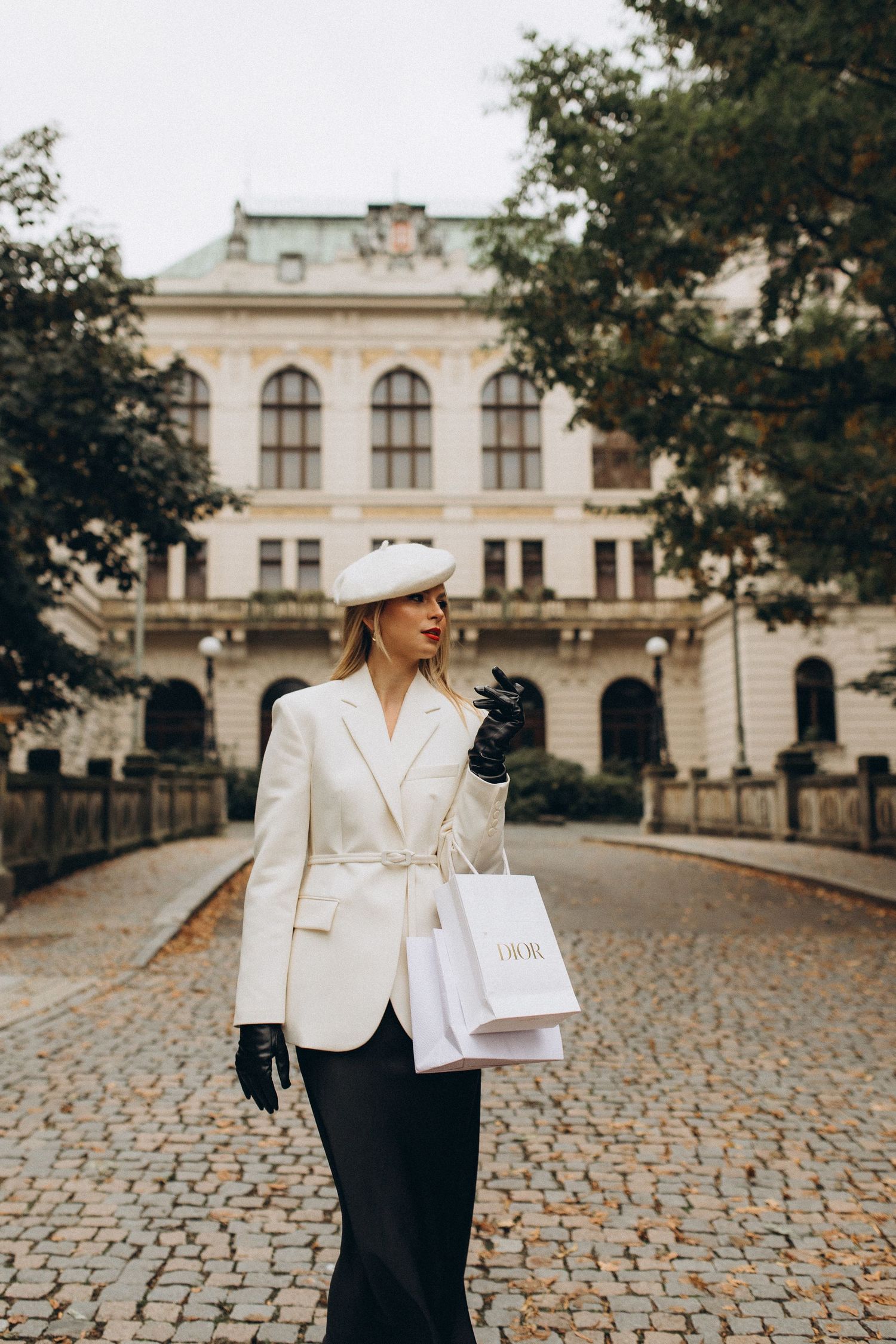 Person wearing white blazer and black skirt walking on cobblestone street near historic building.