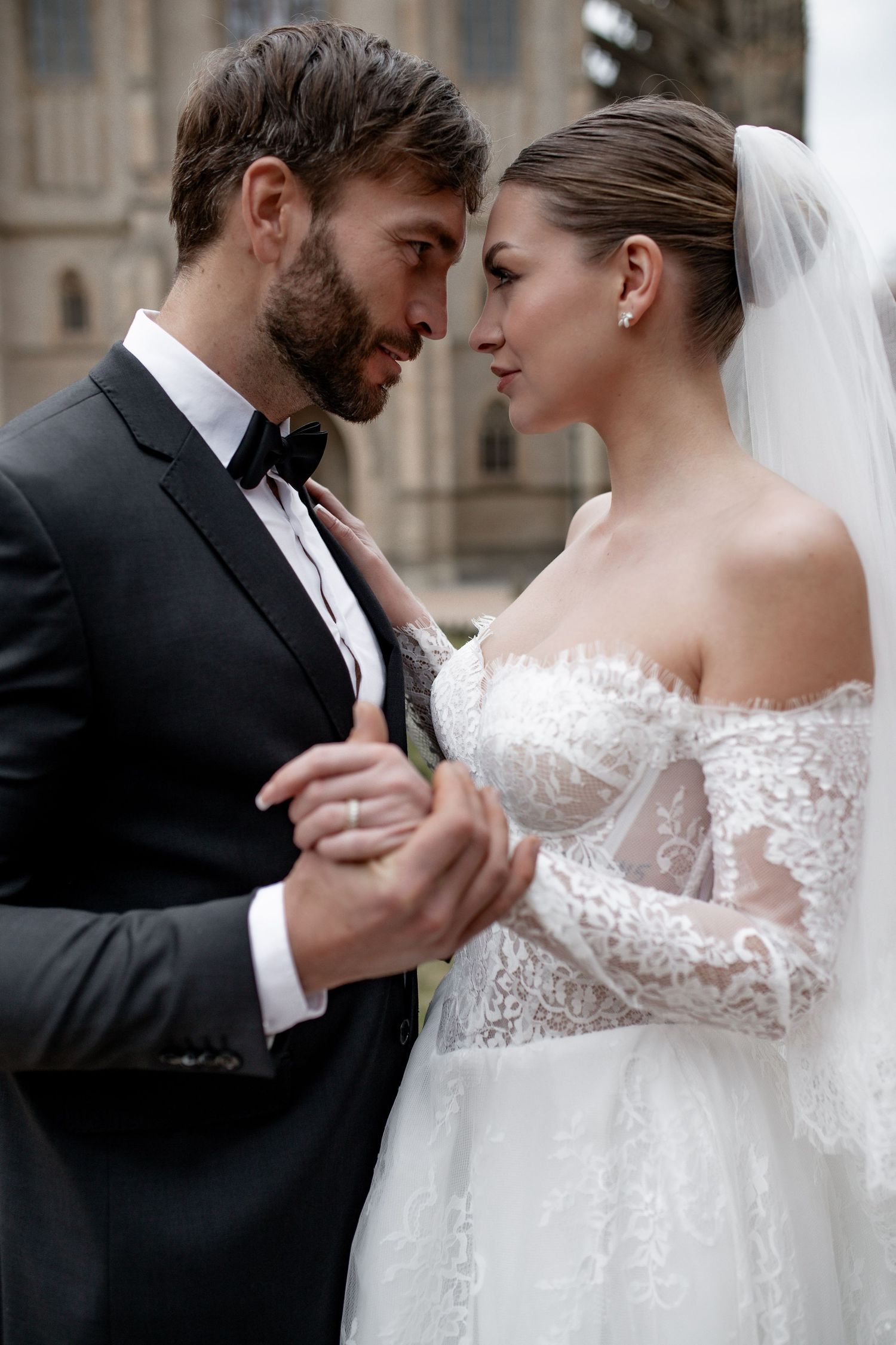 A bride and groom share an intimate moment, touching foreheads, dressed in elegant wedding attire outdoors.