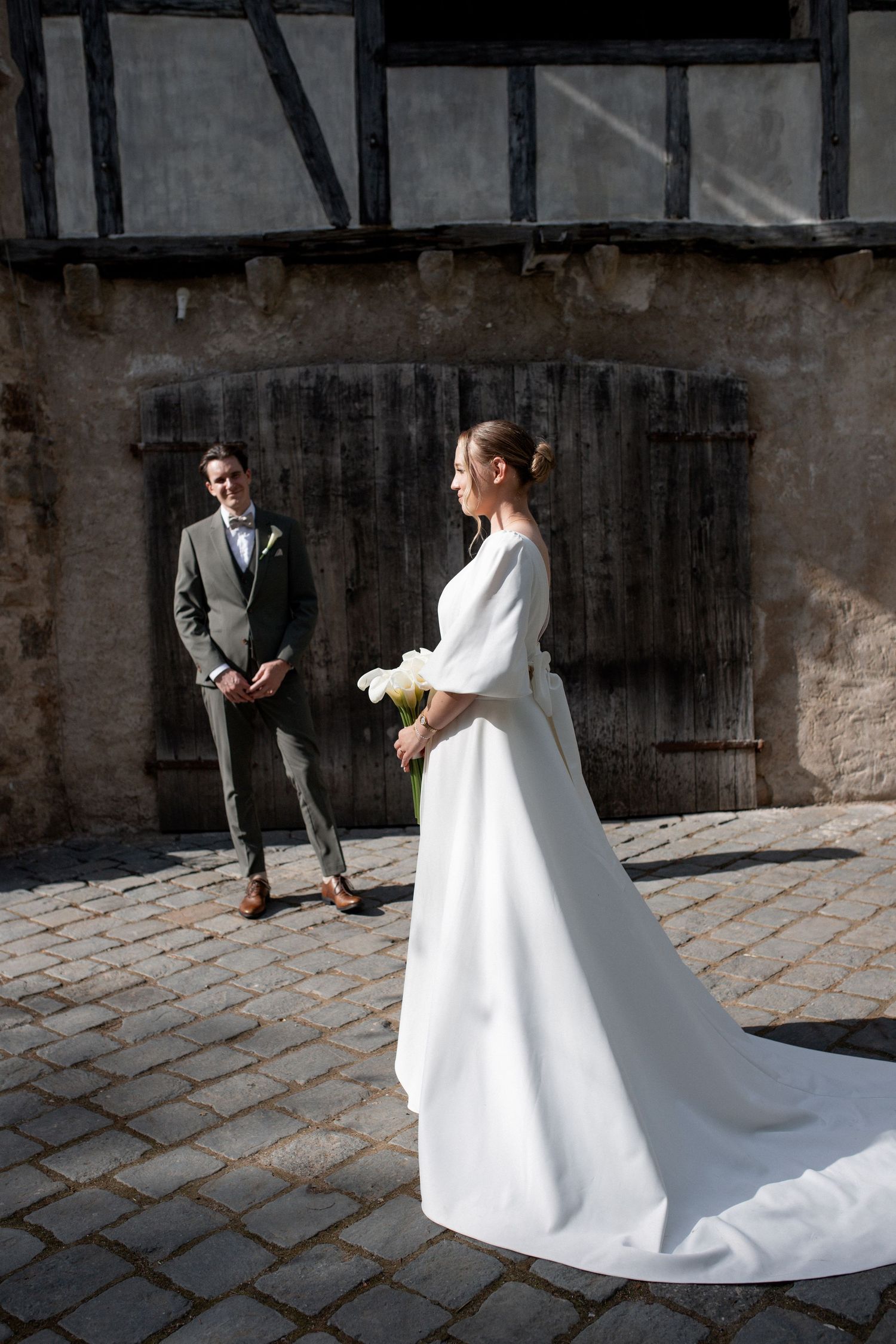 Bride in white gown holds bouquet facing groom in gray suit on cobblestone courtyard near rustic building.