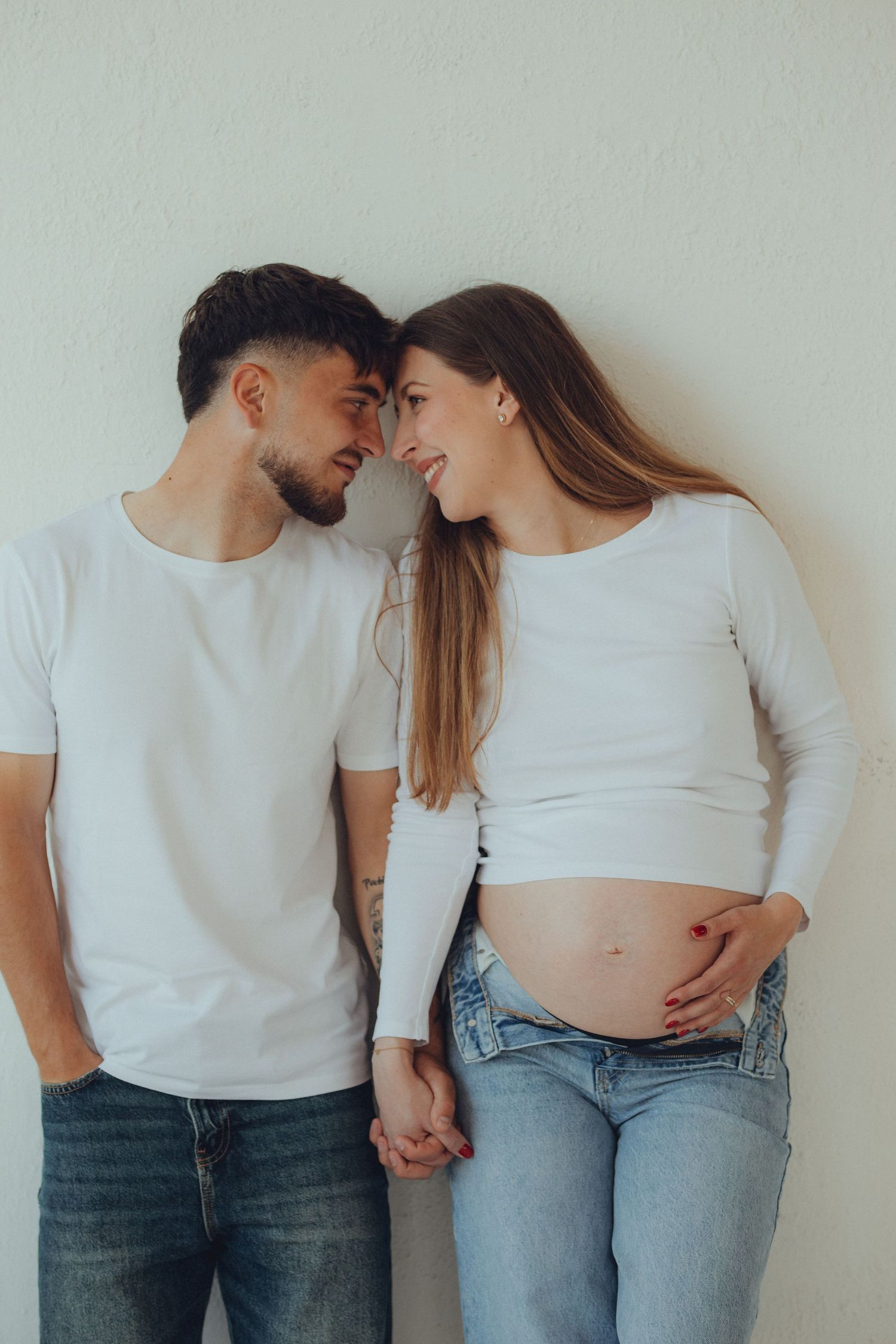 Expecting couple in white tops and jeans share an intimate moment against a light wall while showing her pregnant belly.