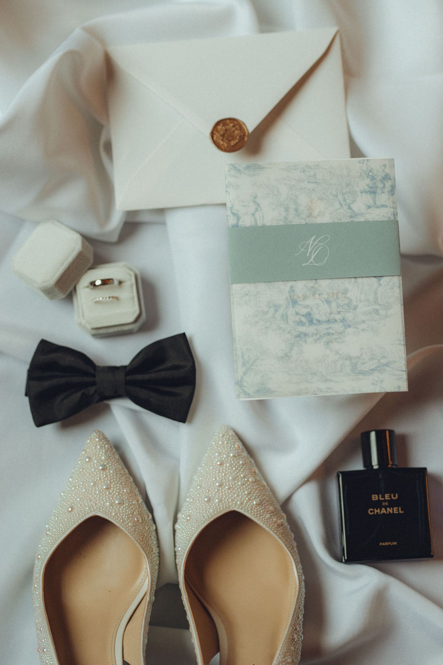Wedding flat lay with pearl heels, bow tie, rings, Bleu de Chanel perfume, invitation, and wax seal envelope on white silk.