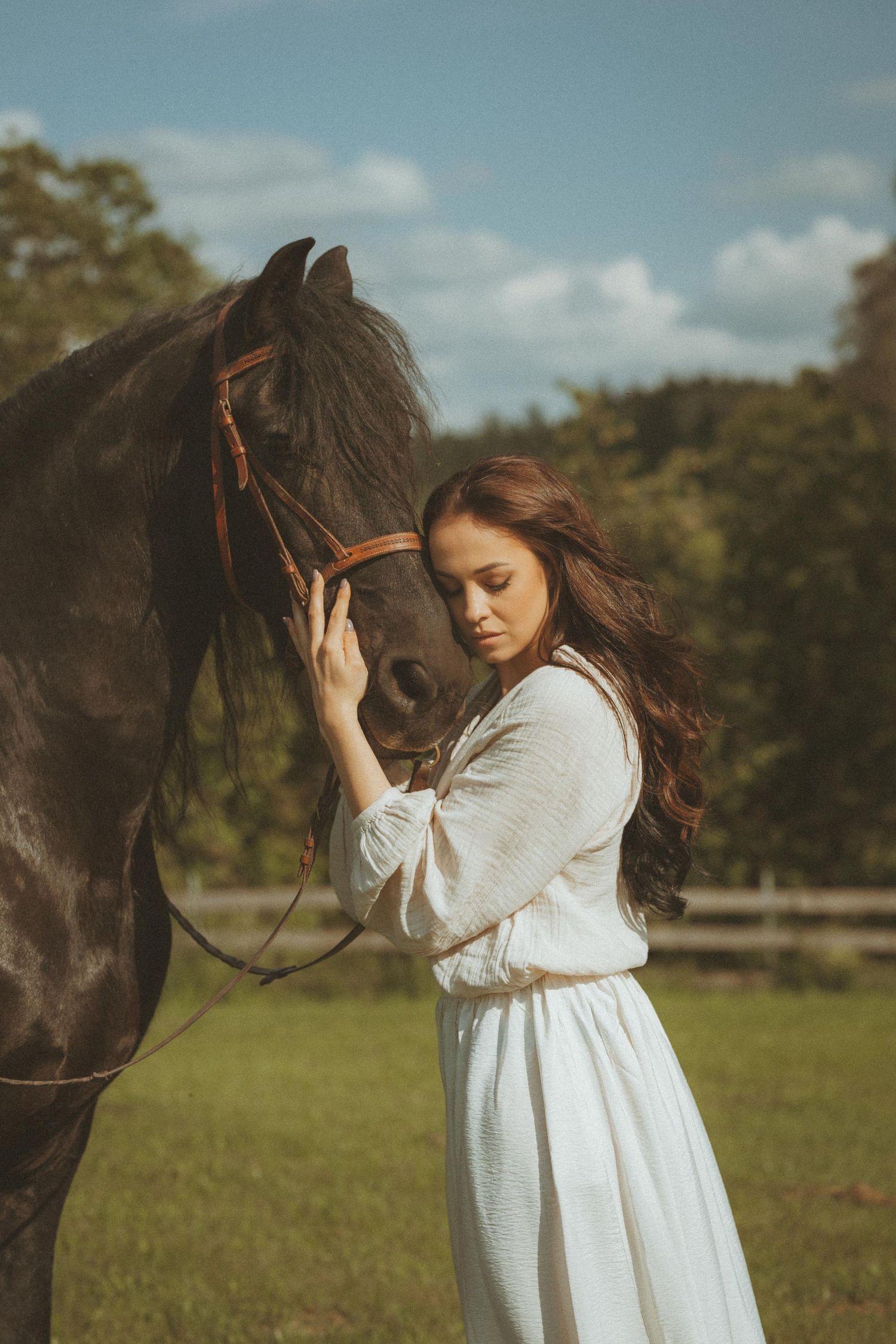 An intimate moment captured as a woman in white dress gently touches a dark horse's face in a pastoral setting.