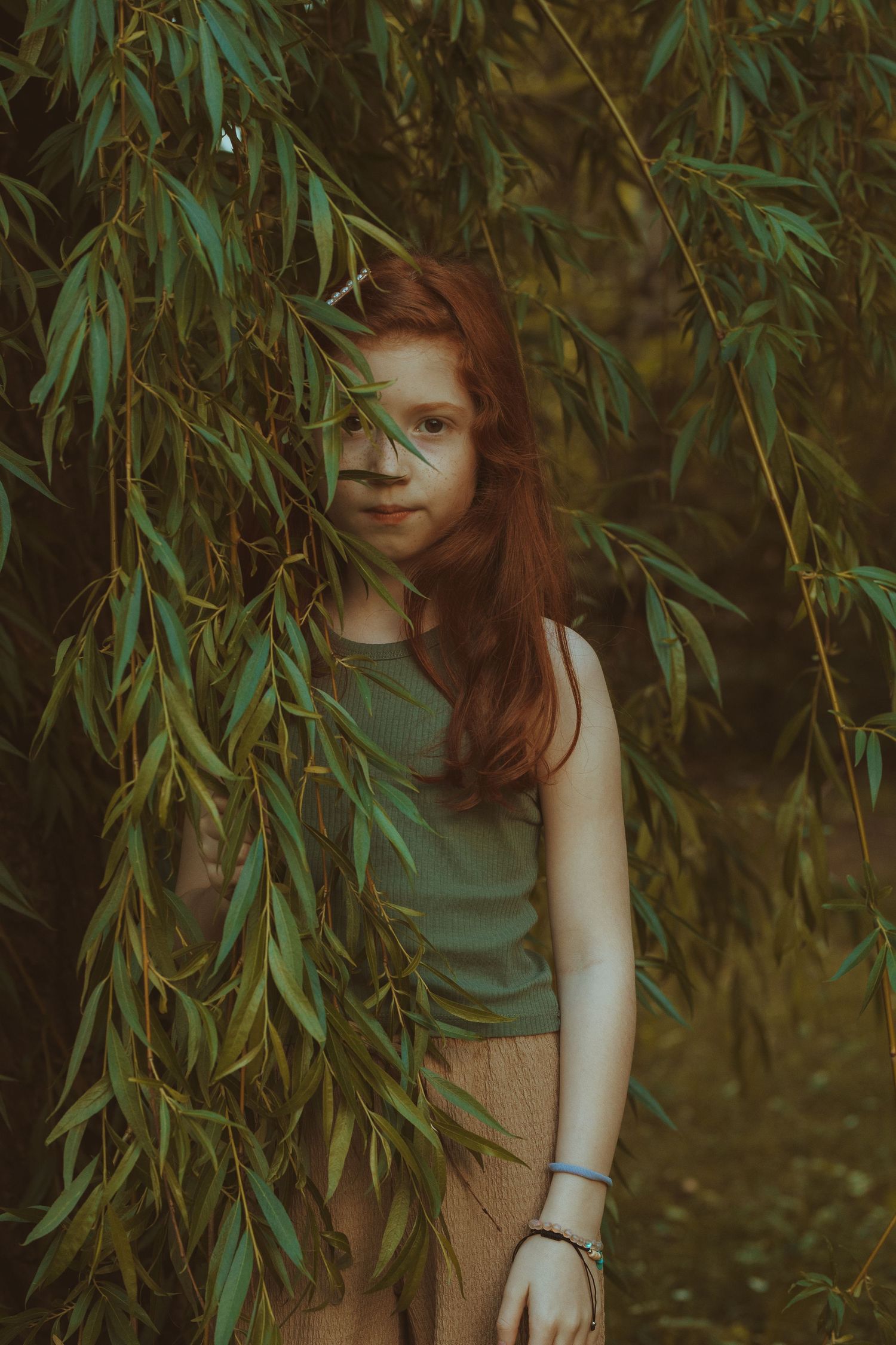 Portrait of a girl in green tank top posing among willow tree branches.