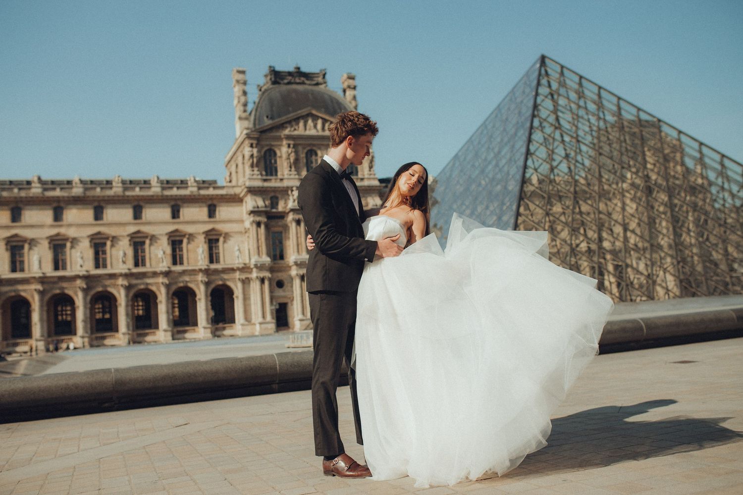 A romantic wedding couple embraces in front of the iconic Louvre Museum and glass pyramid in Paris, France.