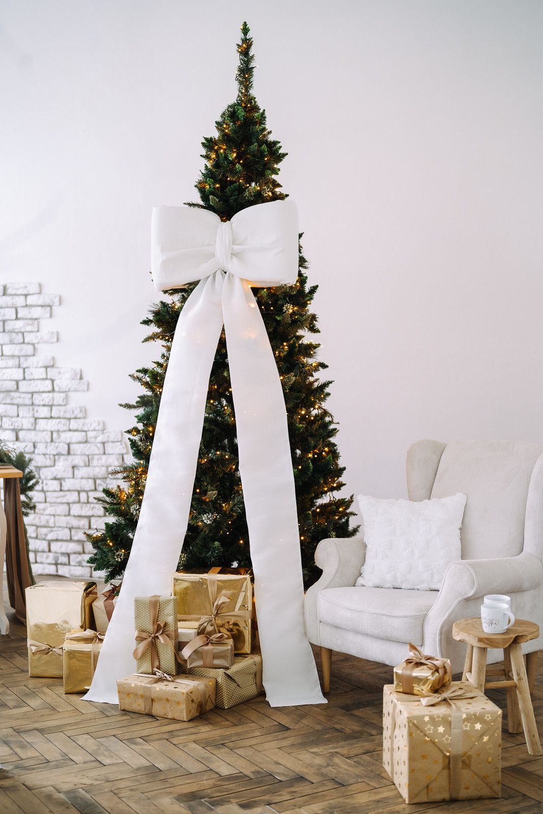 White Christmas decorations with large bows and wrapped gold presents arranged beside evergreen trees against white brick wall.
