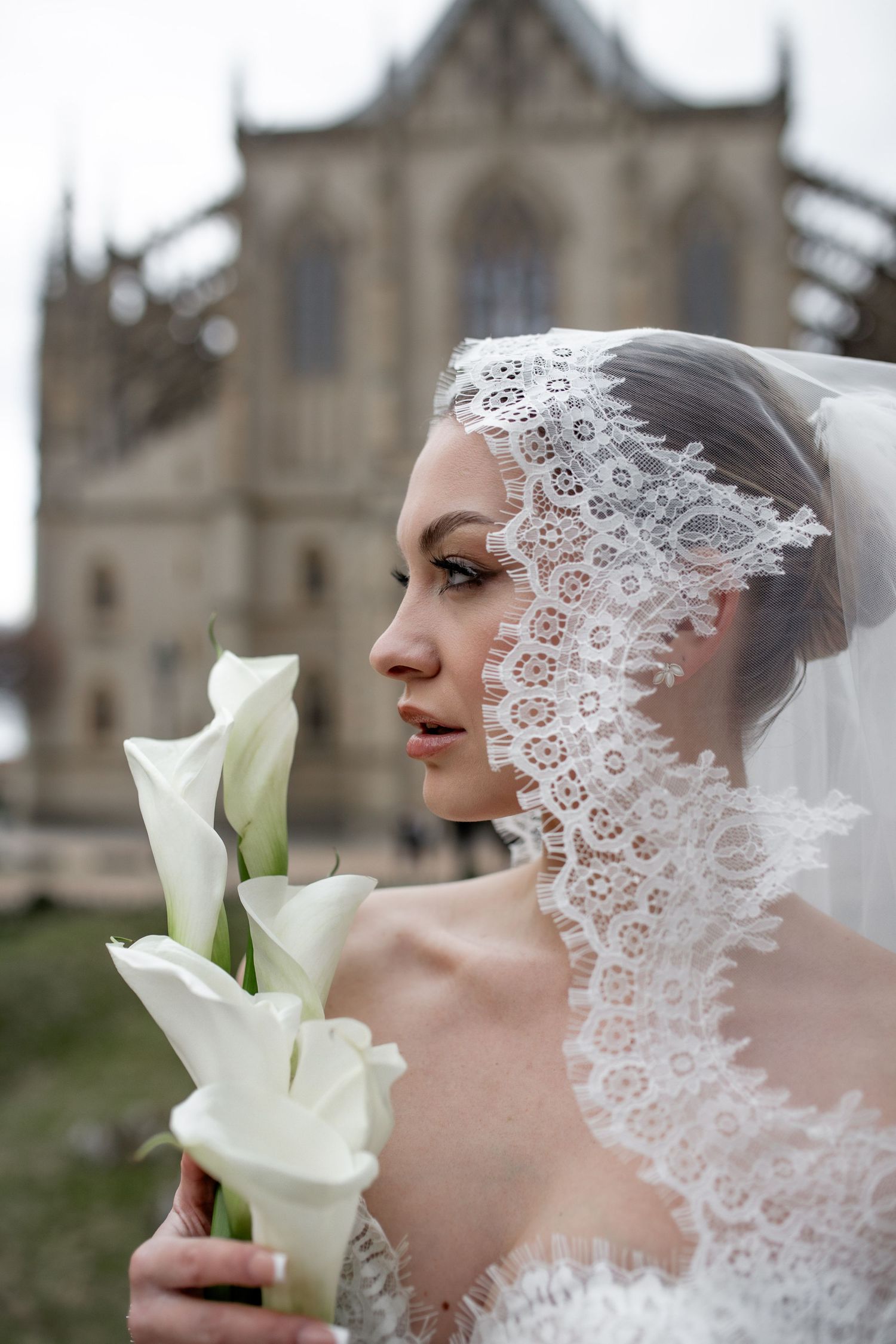 Elegant bride in lace veil holding white calla lilies with gothic church architecture in background.