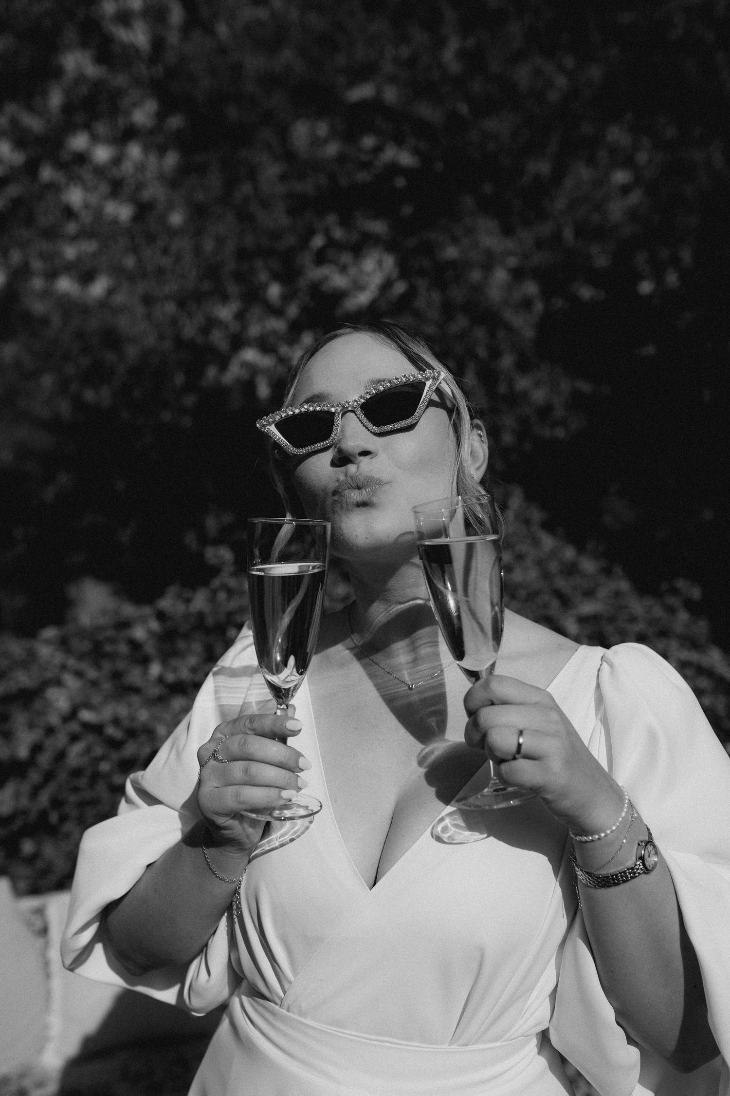 Black and white photo of a bride in sunglasses holding two champagne flutes outdoors.