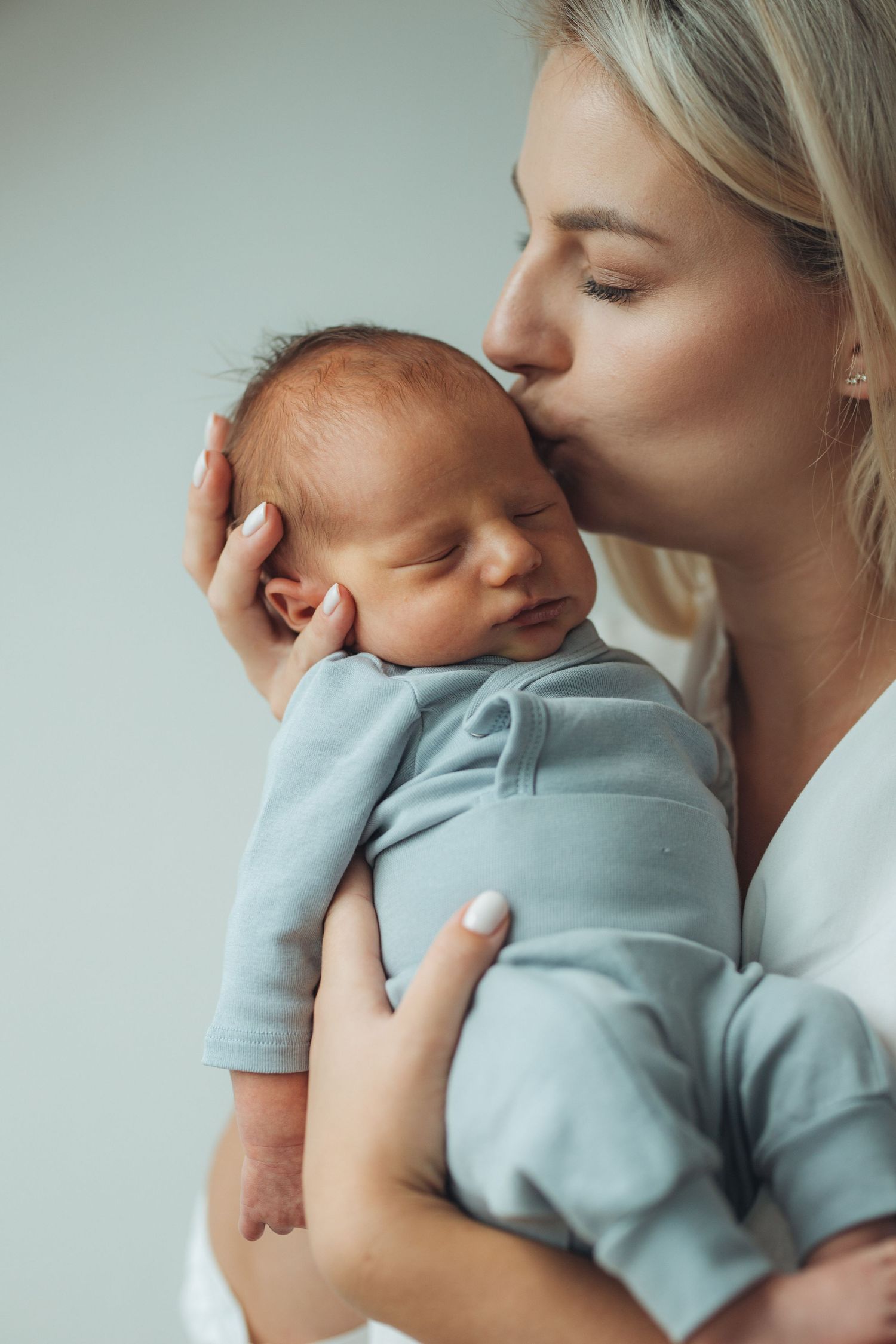 A tender moment as a blonde mother gently kisses her sleeping newborn baby dressed in a soft blue outfit.