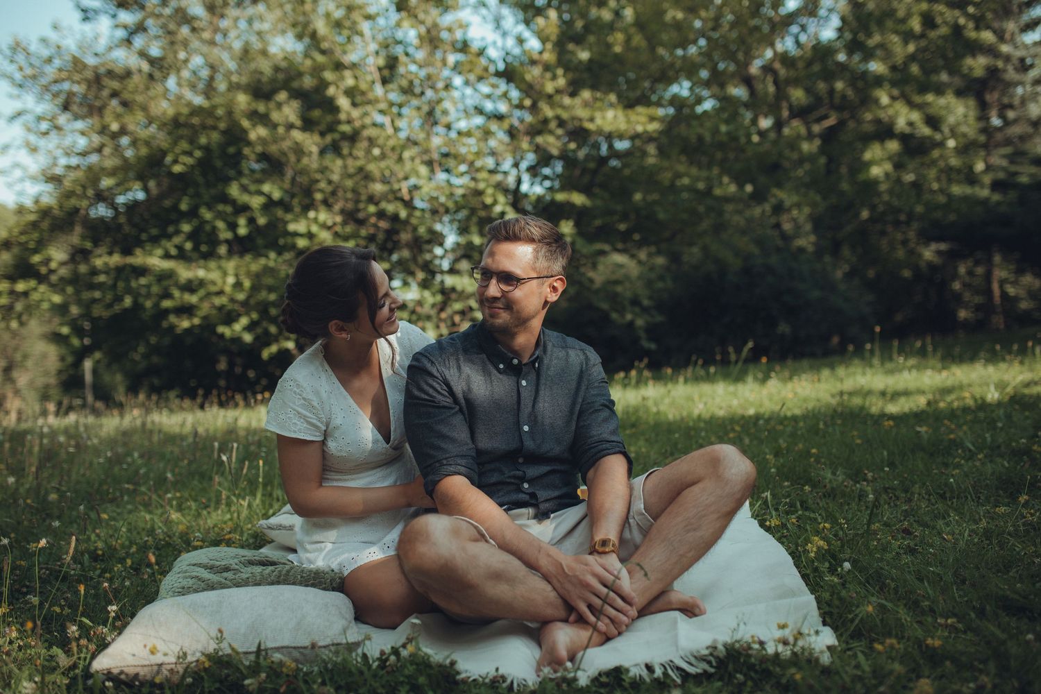 A couple shares an intimate moment while sitting on a blanket in a sunny field.