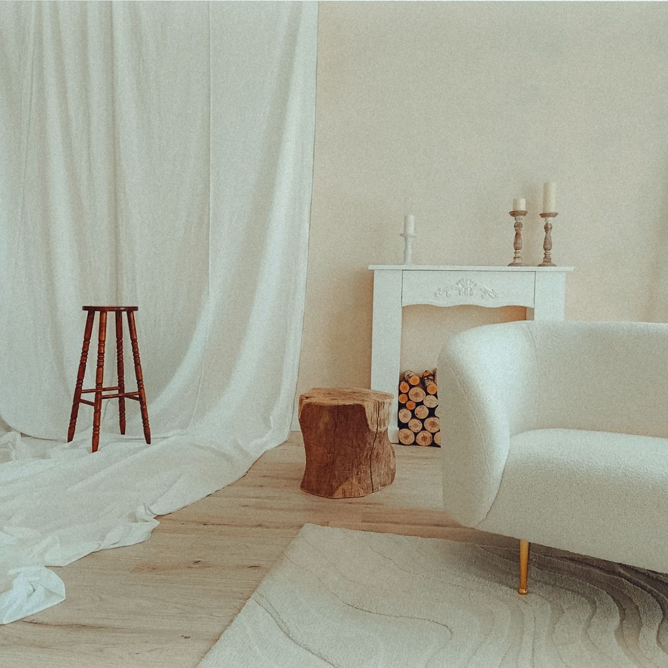 Minimalist living room with white curtains, wooden stool, tree stump side table and modern armchair beside a decorative fireplace.