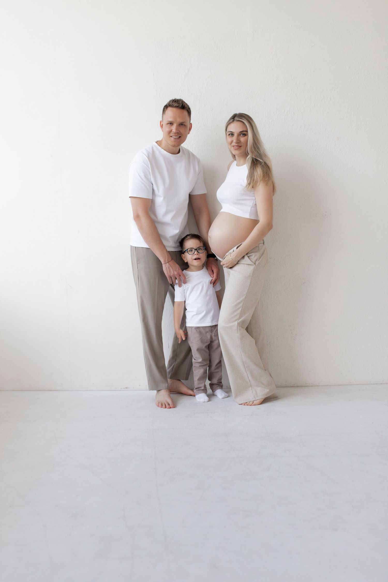 Happy family with pregnant mother, father, and young child in glasses posing together in white studio.