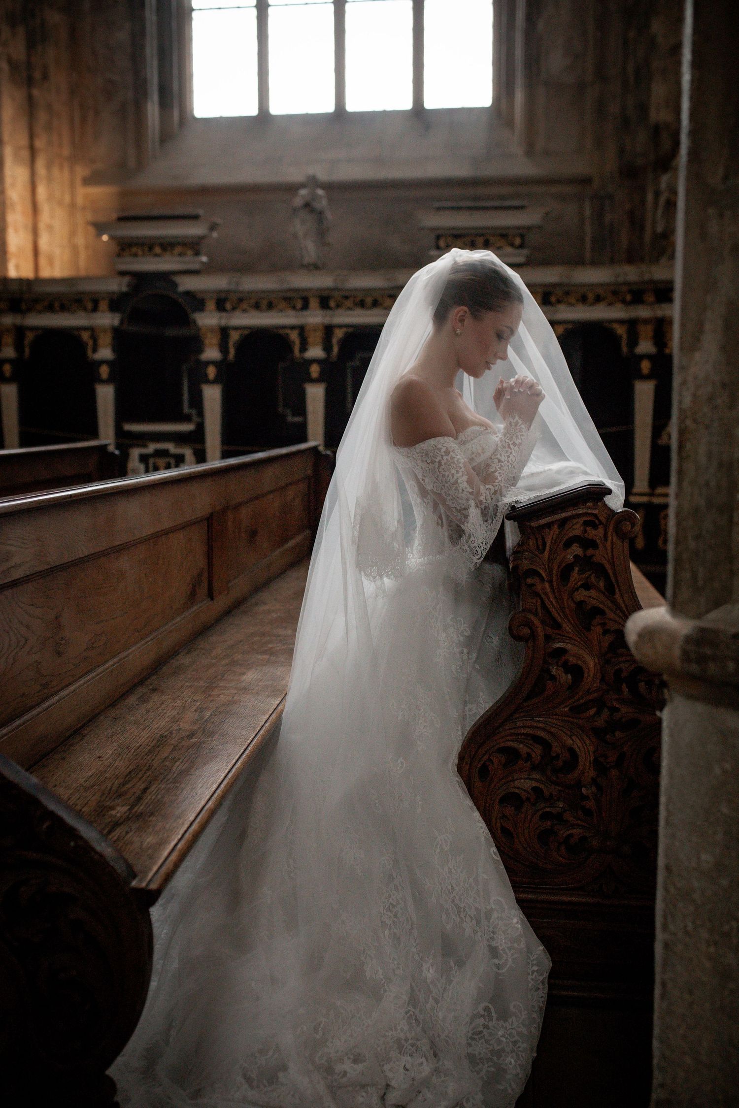 Bride in white lace gown and veil kneeling in prayer at ornate wooden church pew during wedding ceremony.