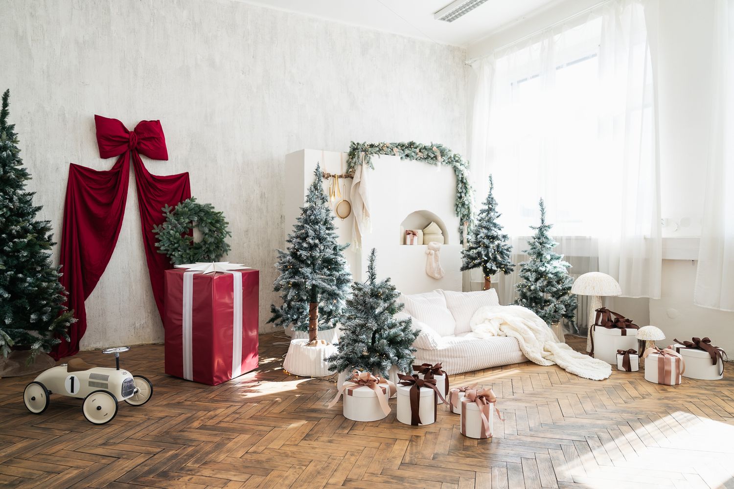 Festive Christmas room with decorated trees, large gift boxes, red bow curtain, and a white couch surrounded by holiday decor.