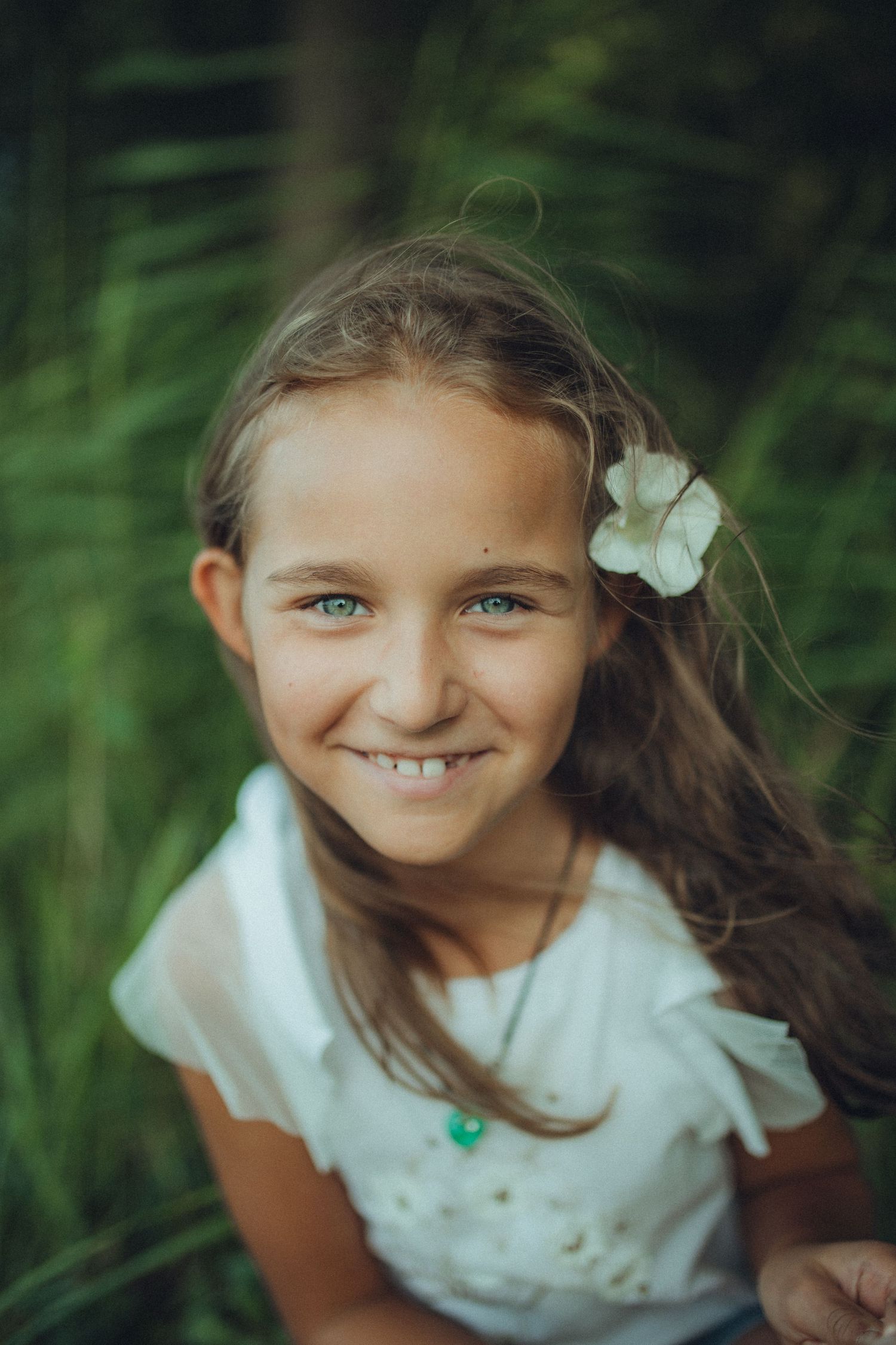 Close-up portrait of a child with a white flower in her hair against green foliage.