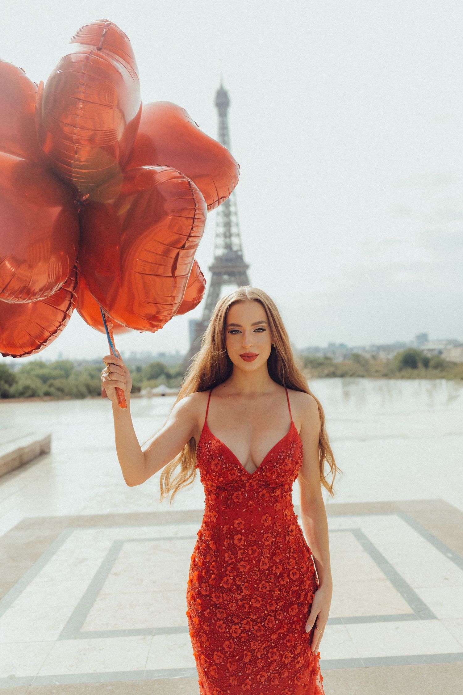 Woman in sparkly red dress holding heart-shaped balloons with Eiffel Tower in background.