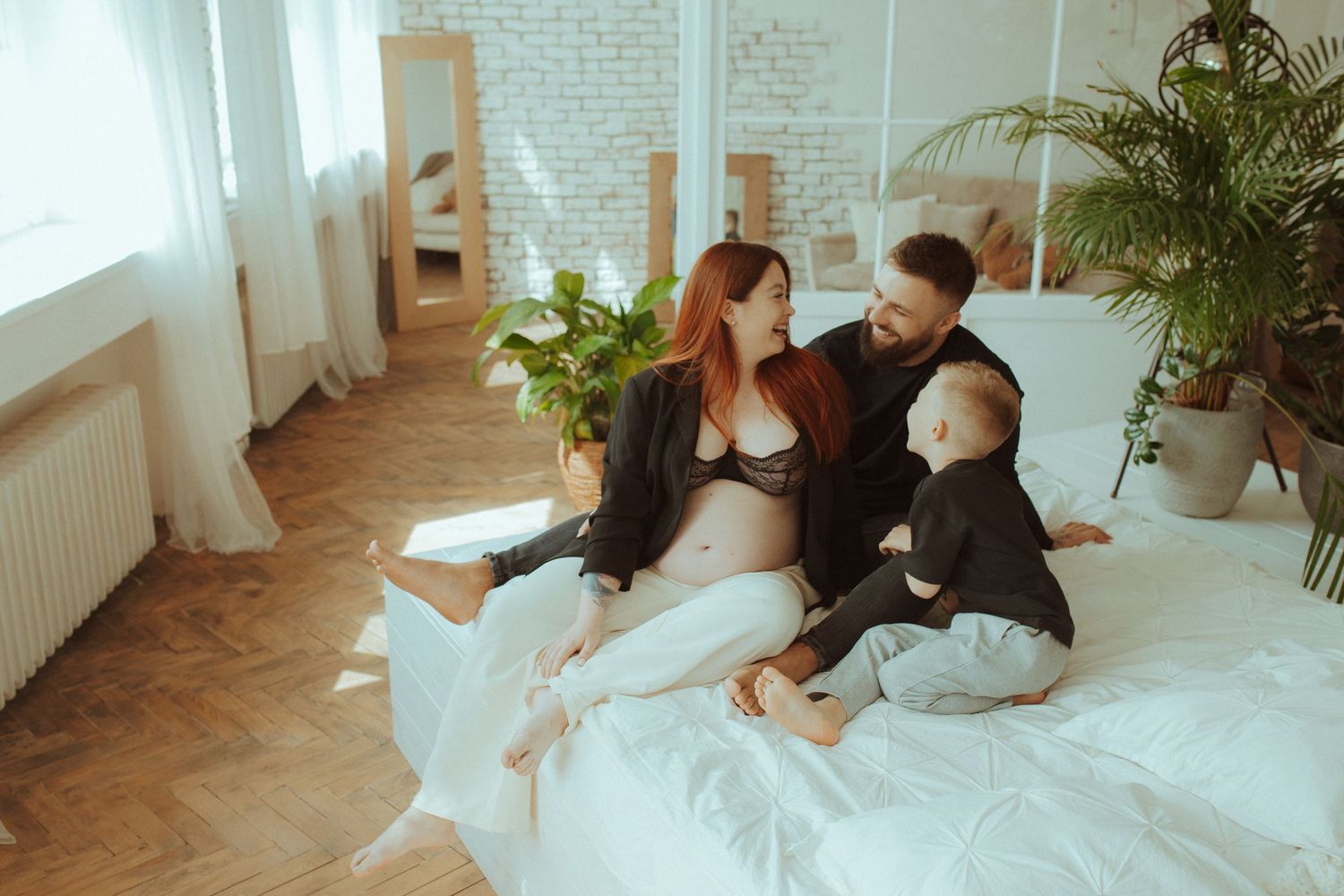 A couple cuddles on a bright white floor near a window with potted plants in the background.