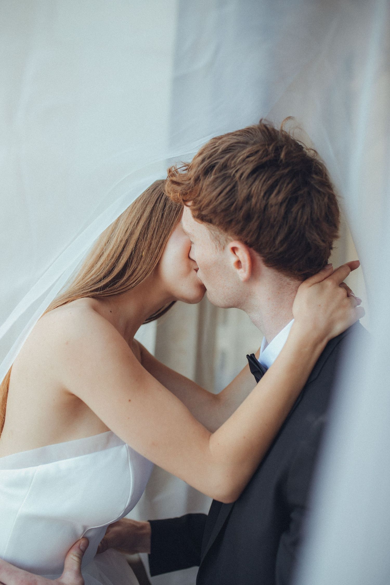 A tender romantic moment between a couple in formal wedding attire sharing a kiss against a light background.