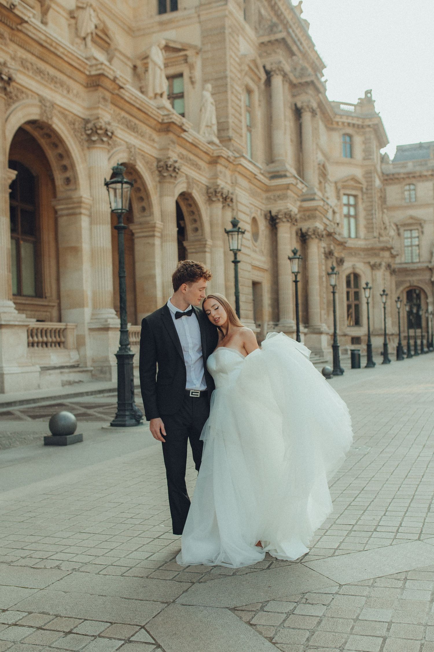 A couple during a wedding photoshoot strolls through an elegant European palace courtyard with ornate architecture.