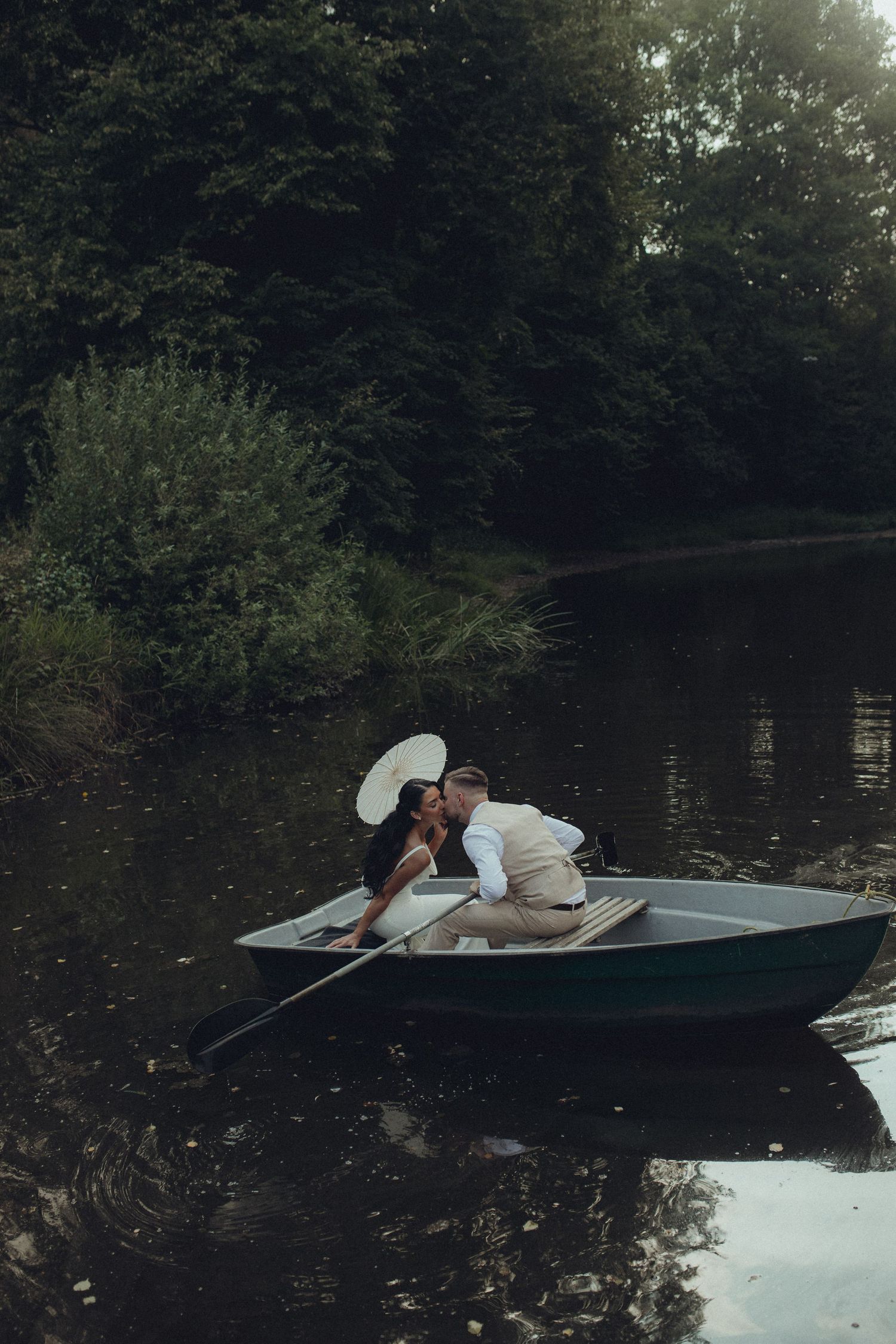 A couple shares a romantic kiss in a rowboat on a calm lake surrounded by lush green trees.