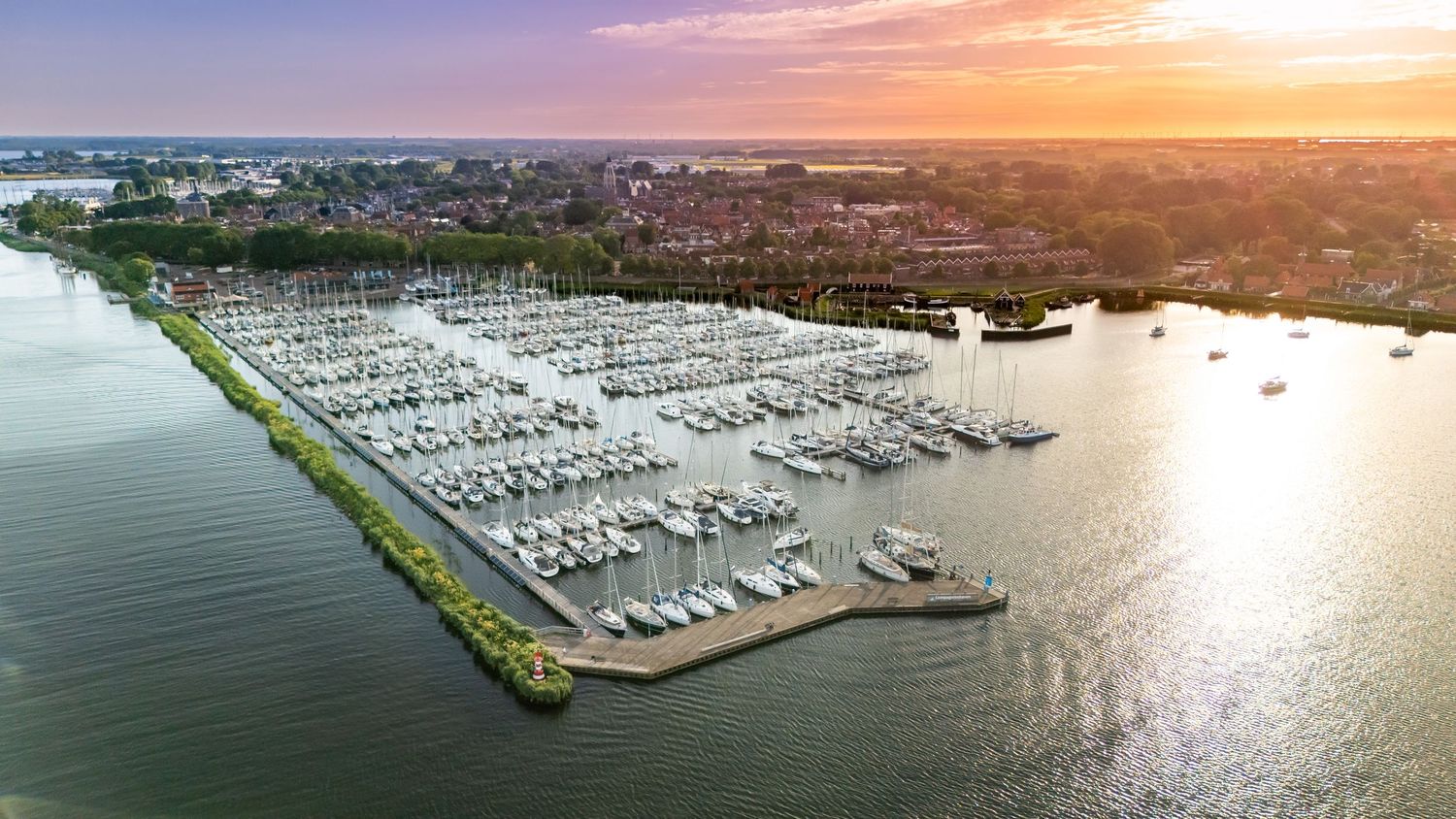Aerial sunset view of a busy marina with white sailboats docked along piers, surrounded by calm water and waterfront homes.