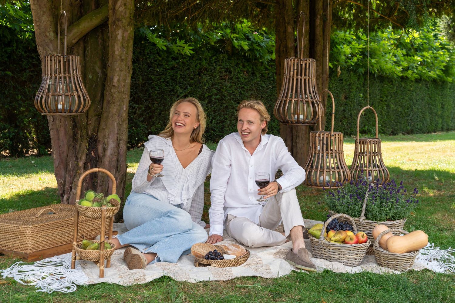 Couple enjoying a romantic outdoor picnic with red wine, fruit baskets, and hanging lanterns on a sunny garden lawn.