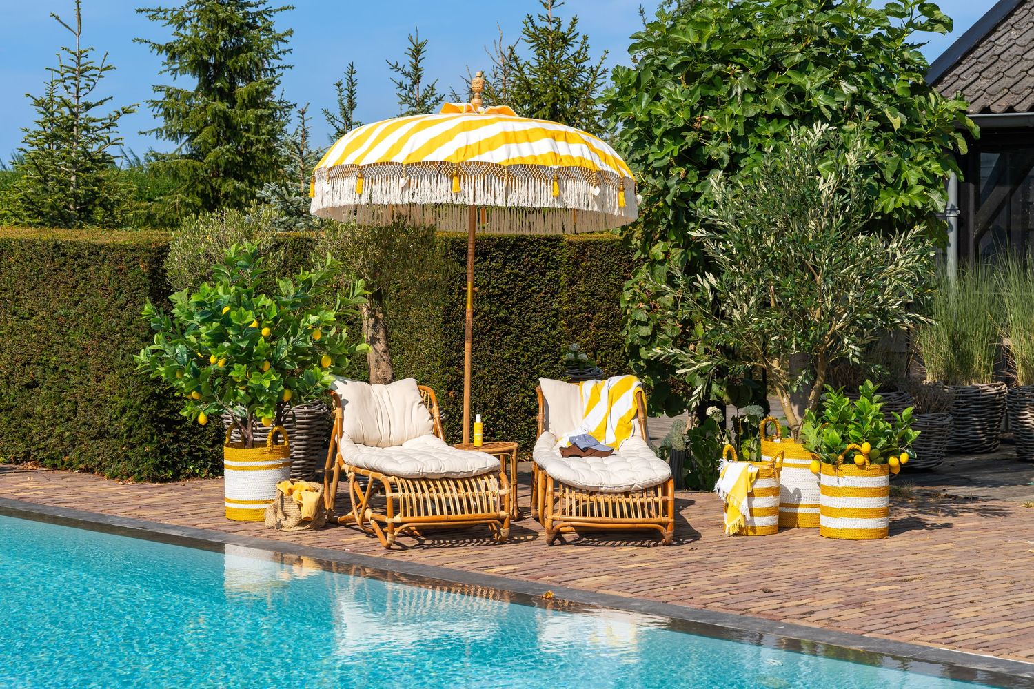 Yellow striped patio umbrella with fringe over rattan chairs beside a pool, surrounded by lush greenery and potted plants.