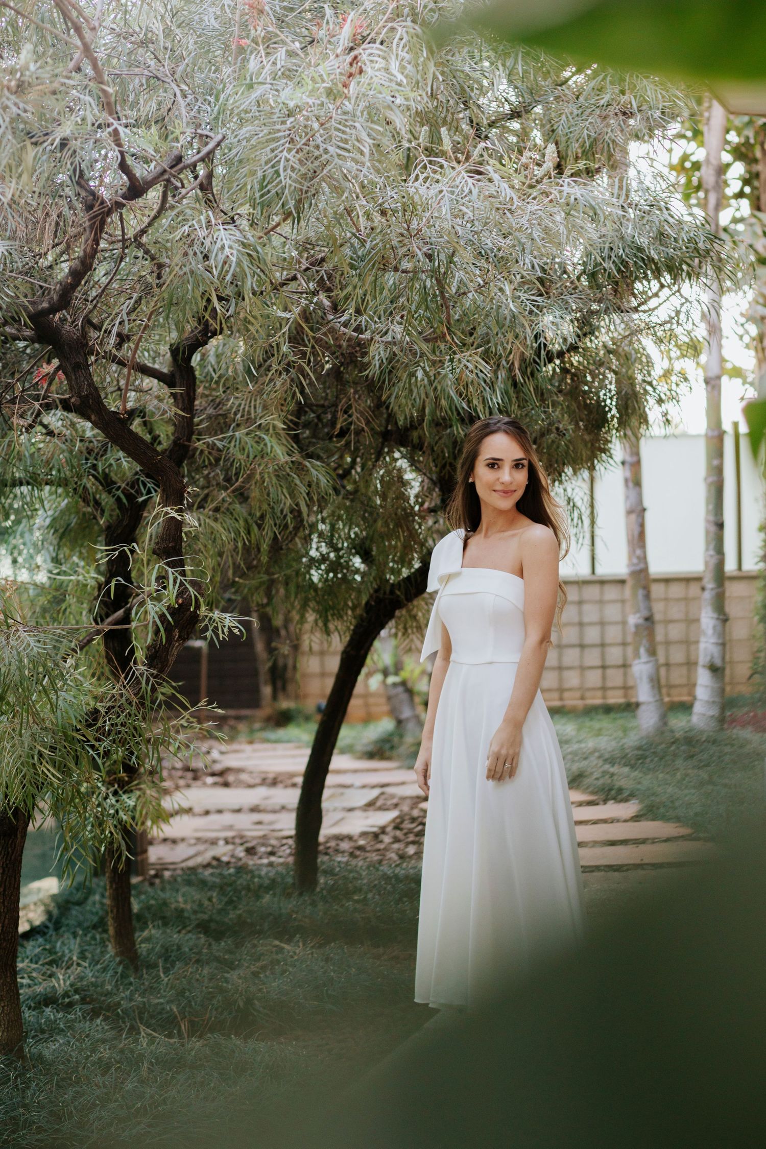 A bride in a flowing white dress poses elegantly among olive trees in a garden setting during a wedding photoshoot.