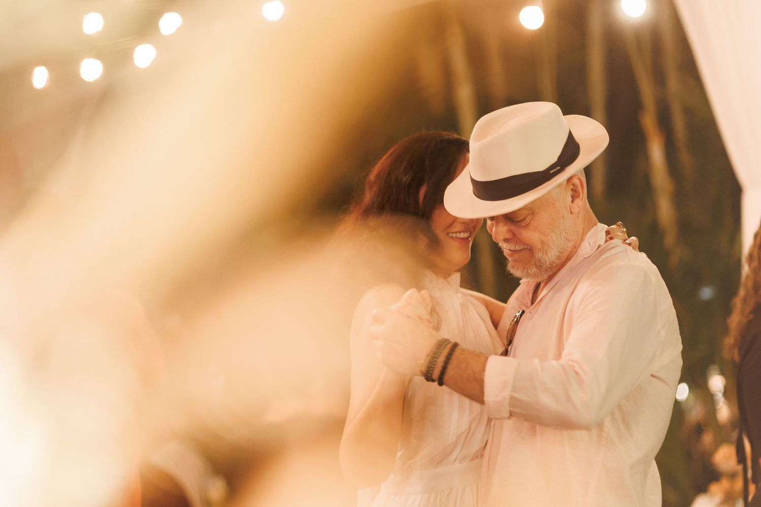 Couple sharing a romantic dance under warm string lights, man wearing white hat and linen shirt at an outdoor evening event.