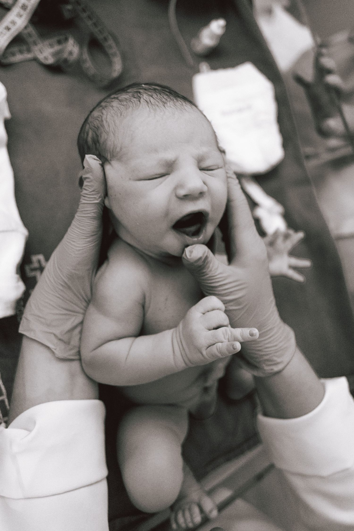 Newborn baby yawning moments after birth, held by gloved medical hands in black and white photo.