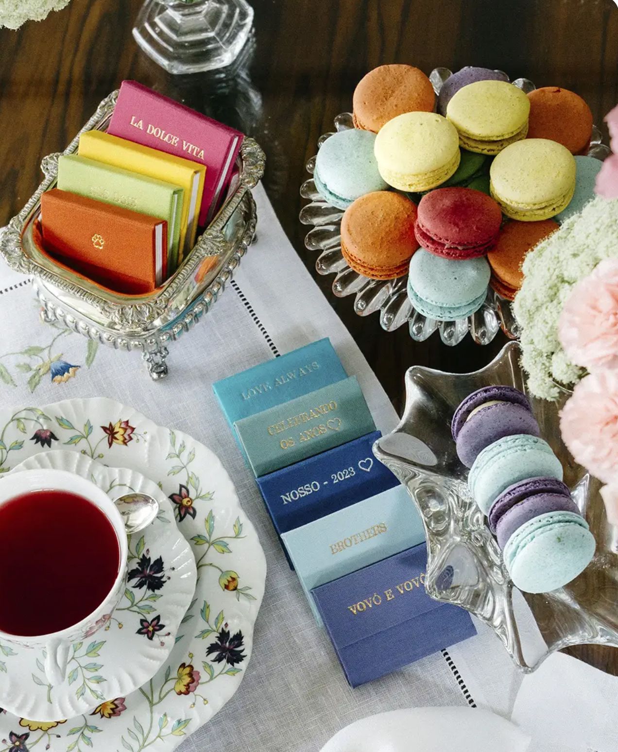Elegant tea party spread with colorful French macarons, matchbooks, floral china teacup, and pastel booklets on a white tablecloth.