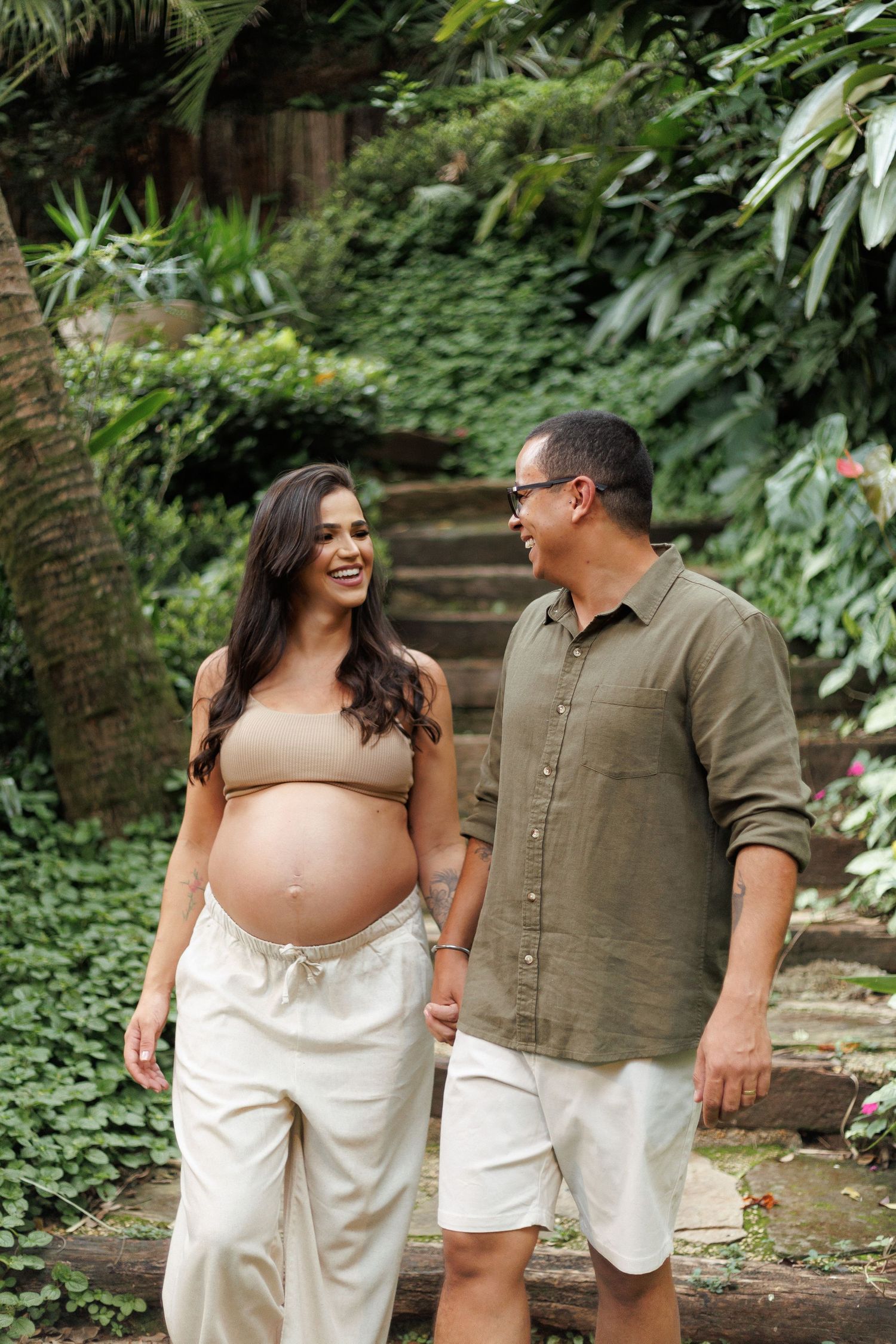 Happy pregnant woman and partner holding hands while walking through a lush tropical garden setting.