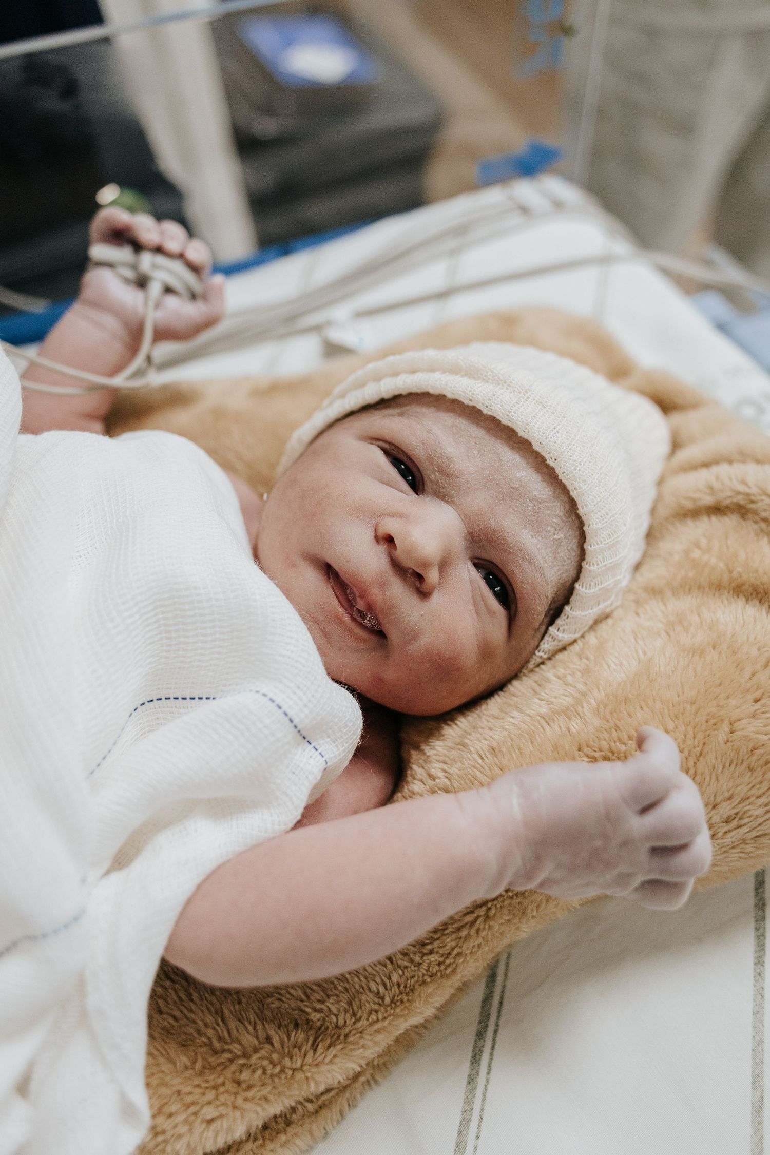 Newborn baby in white cap smiles while resting on soft beige hospital blanket.