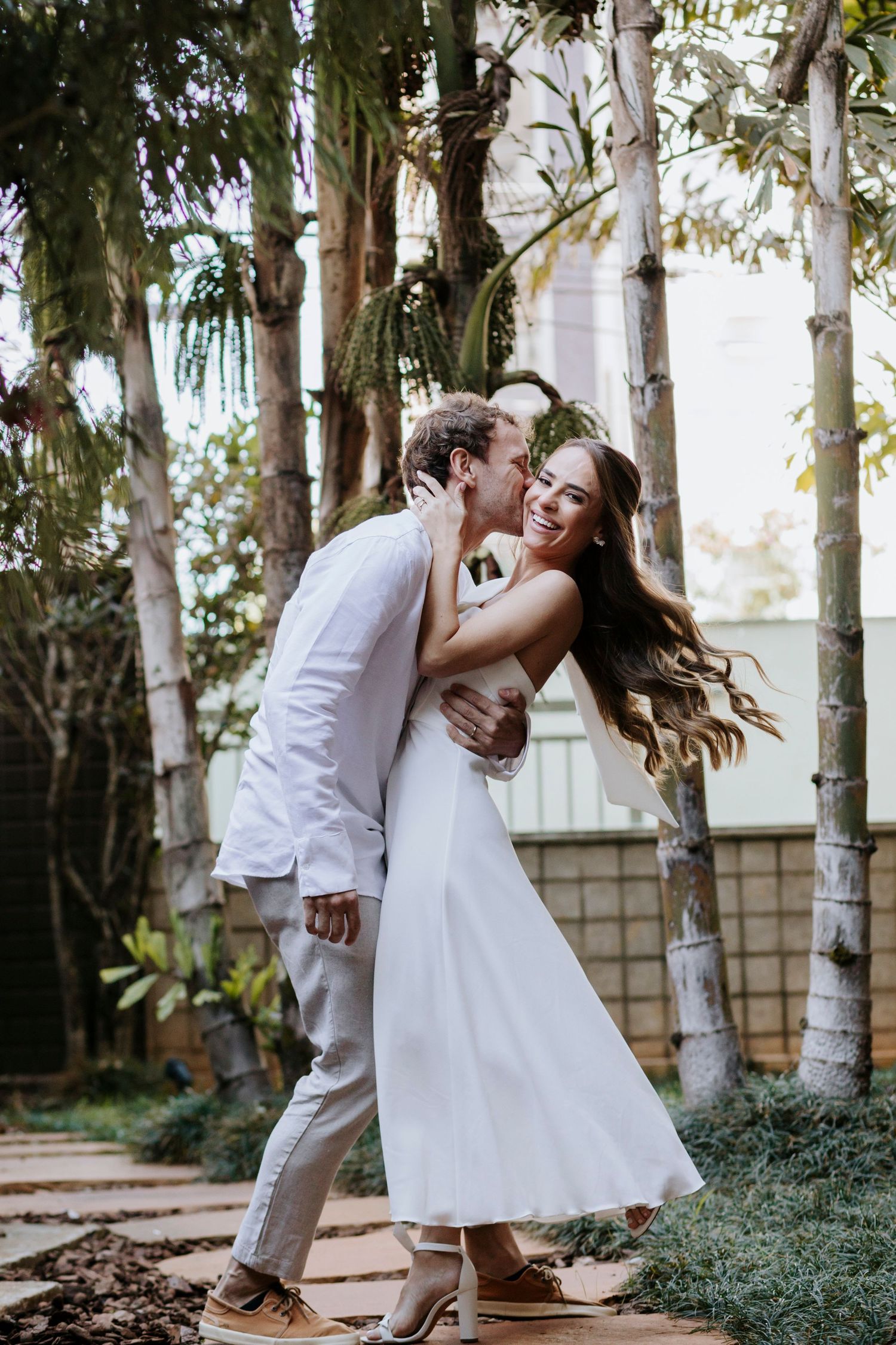 Couple in white attire sharing playful moment among birch trees in garden setting.